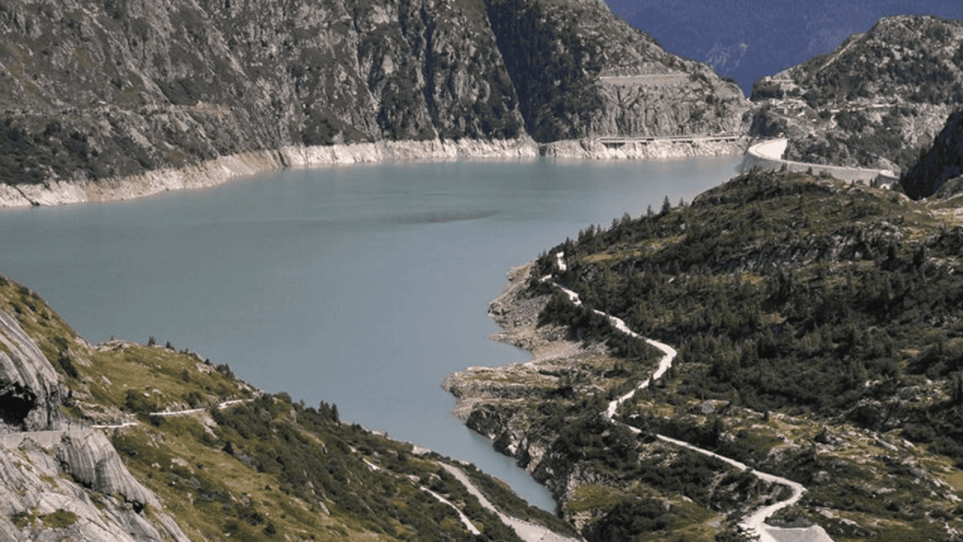 A calm blue lake surrounded by steep, rocky mountains with green vegetation, and a winding road running along the shoreline and up the hillside.