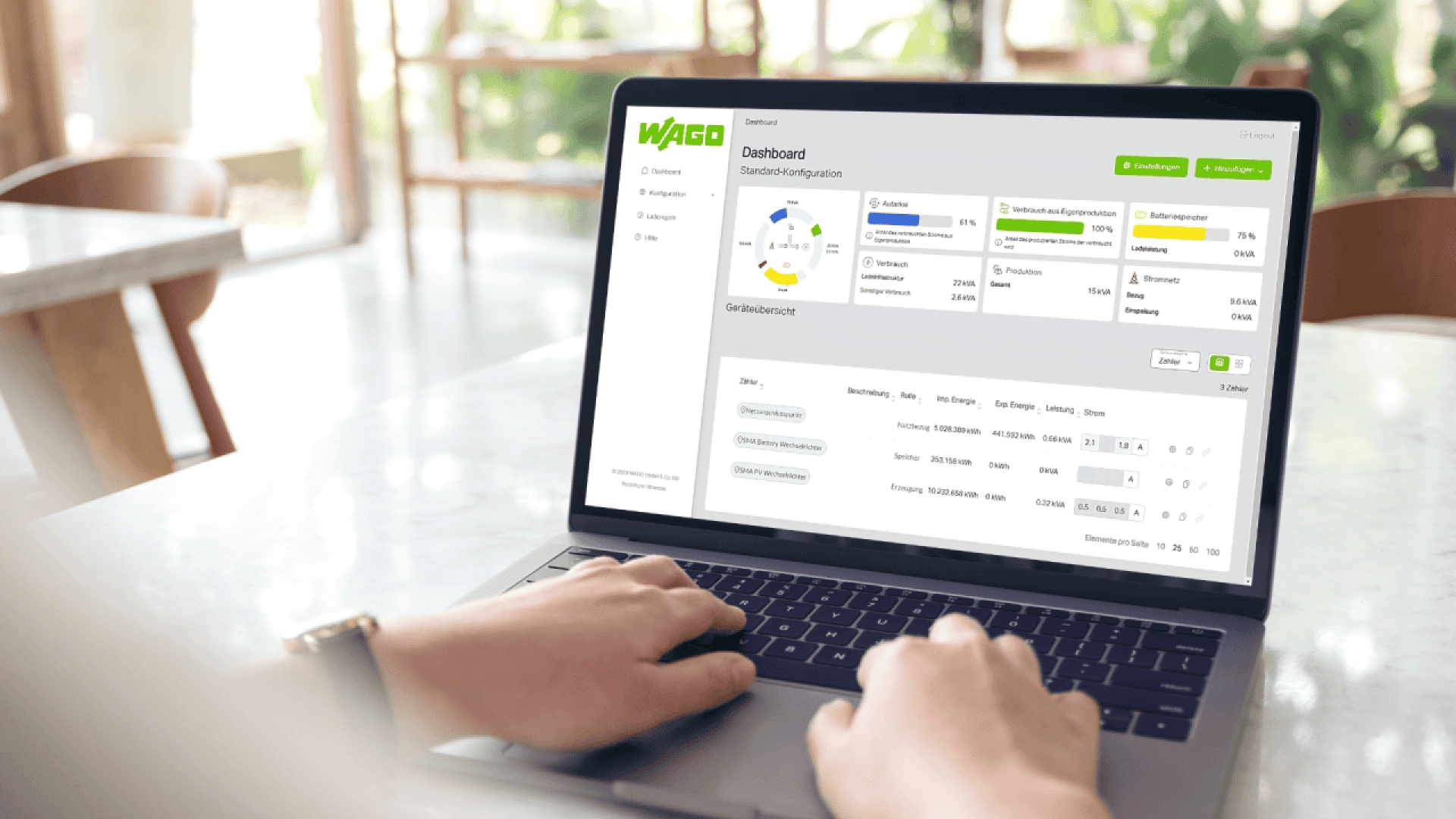 A person uses a laptop displaying a WAGO dashboard with charts, analytics, and configuration options, sitting at a table in a bright room with large windows and green plants in the background.