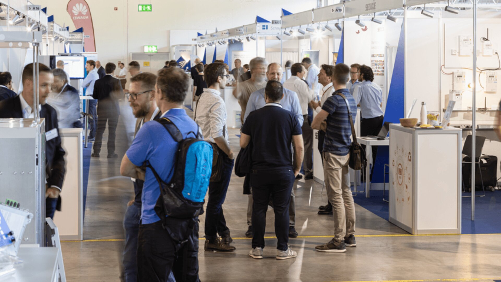 Die Menschen gehen und stehen in Gruppen und unterhalten sich auf einer überdachten Messe oder Ausstellung mit Ständen auf jeder Seite. Einige Besucher tragen Rucksäcke und Freizeitkleidung, andere sind eher formell gekleidet. Der Raum ist belebt und gut beleuchtet.