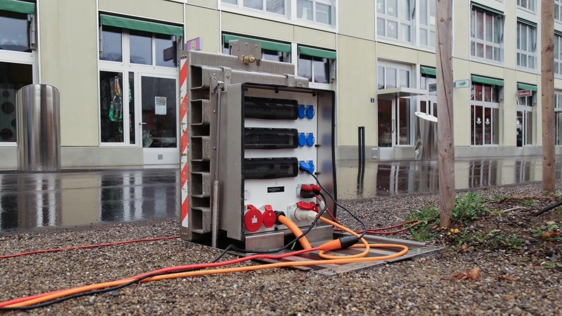 Ein tragbarer Stromverteilerkasten mit mehreren eingesteckten Kabeln steht an einem regnerischen Tag im Freien auf Kies in der Nähe eines Gebäudes mit großen Fenstern.