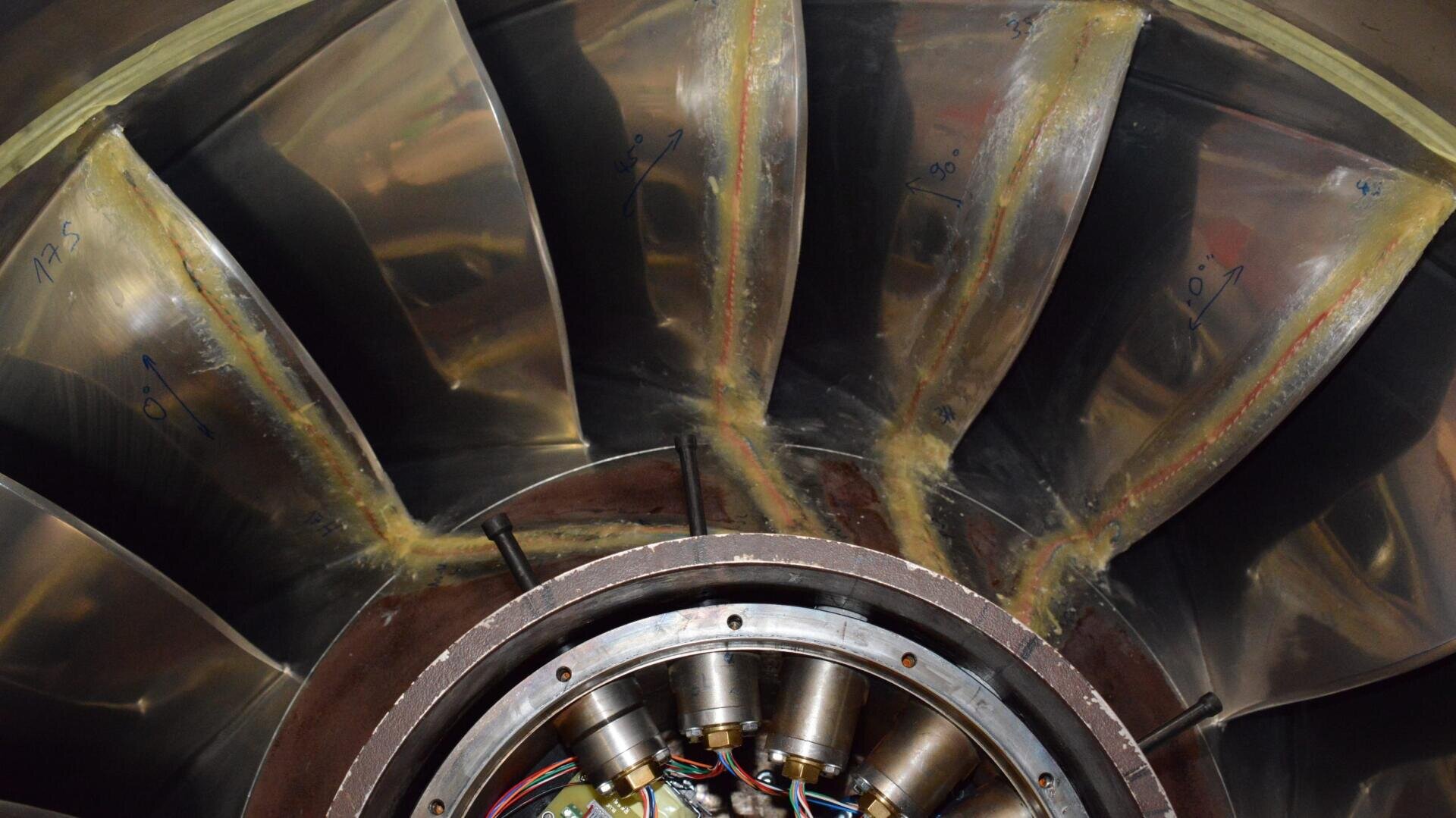 Nahaufnahme des Innenraums einer Metallturbine oder eines Düsentriebwerks, mit glänzenden, gebogenen Schaufeln und sichtbaren Kabeln und Bauteilen in der Mitte. Auf mehreren Schaufeln sind handschriftliche Nummern angebracht.