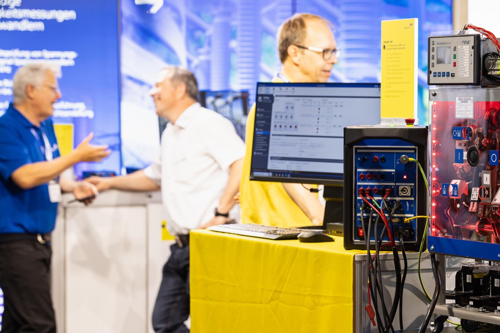 Ein Computerarbeitsplatz mit technischen Geräten steht auf einem gelben Tisch im Vordergrund, während sich drei Männer im unscharfen Hintergrund auf einer Technologiemesse oder -konferenz unterhalten.