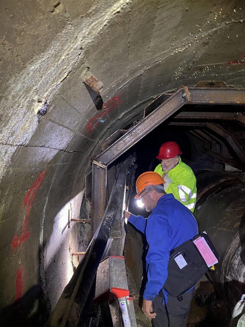 Zwei Arbeiter mit Schutzhelmen und Sicherheitsausrüstung inspizieren einen mit Beton ausgekleideten Tunnel. Einer leuchtet mit einer Taschenlampe auf eine Metallstütze, während der andere beobachtet und sich Notizen macht. An den Tunnelwänden sind rote Markierungen zu sehen.