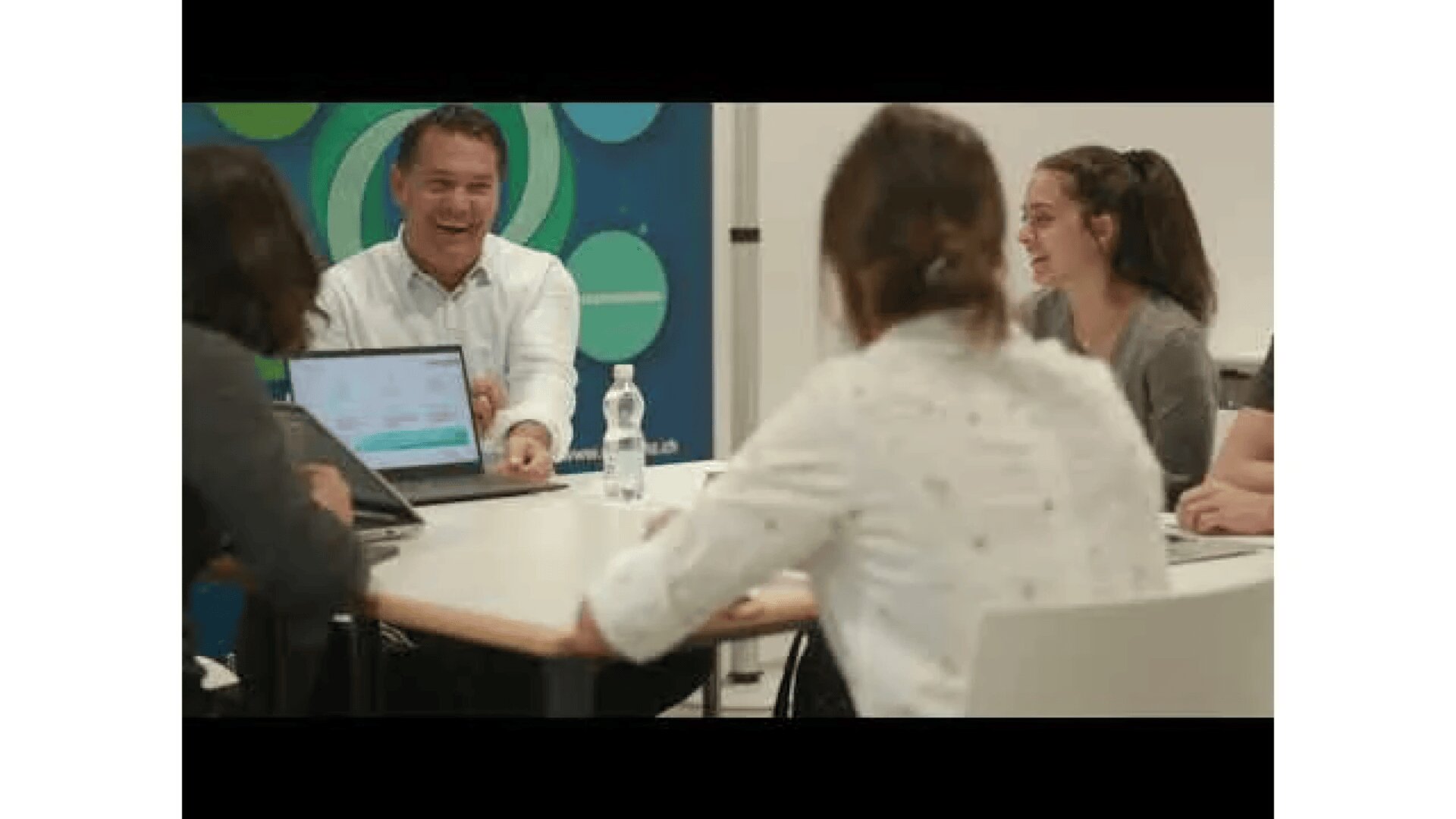 Vier Personen sitzen um einen Tisch herum und diskutieren, wobei ein Mann lächelt und lacht. Auf dem Tisch stehen ein Laptop und eine Wasserflasche, und im Hintergrund ist eine bunte blau-grüne Wand zu sehen.