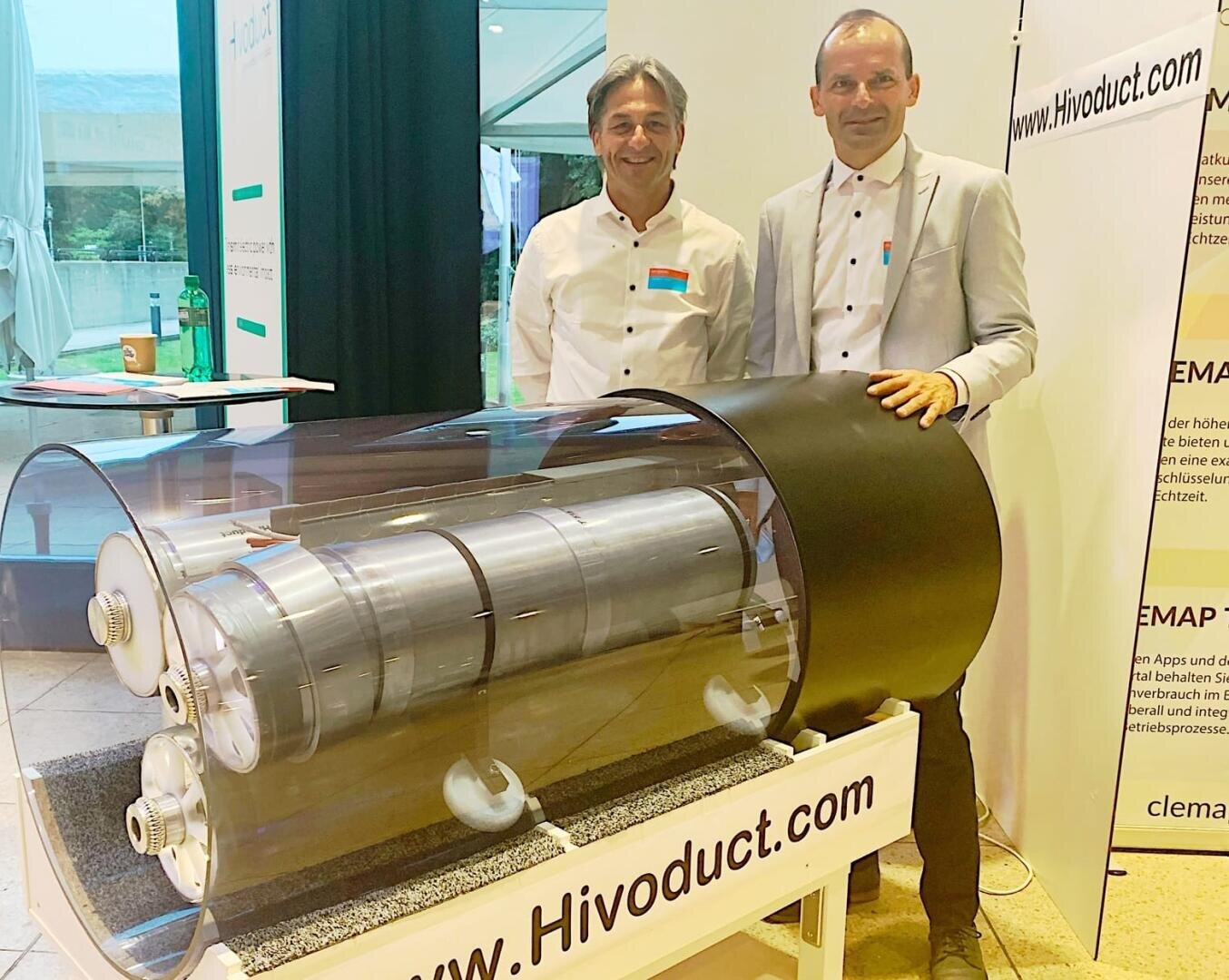 Zwei Männer stehen hinter einem großen zylindrischen Metallgerät, das von durchsichtigem Plastik umhüllt ist und auf einem Tisch mit der Aufschrift "www.Hivoduct.com" bei einer Veranstaltung oder Ausstellung in einem Gebäude ausgestellt ist. Im Hintergrund sind Banner und Fenster zu sehen.