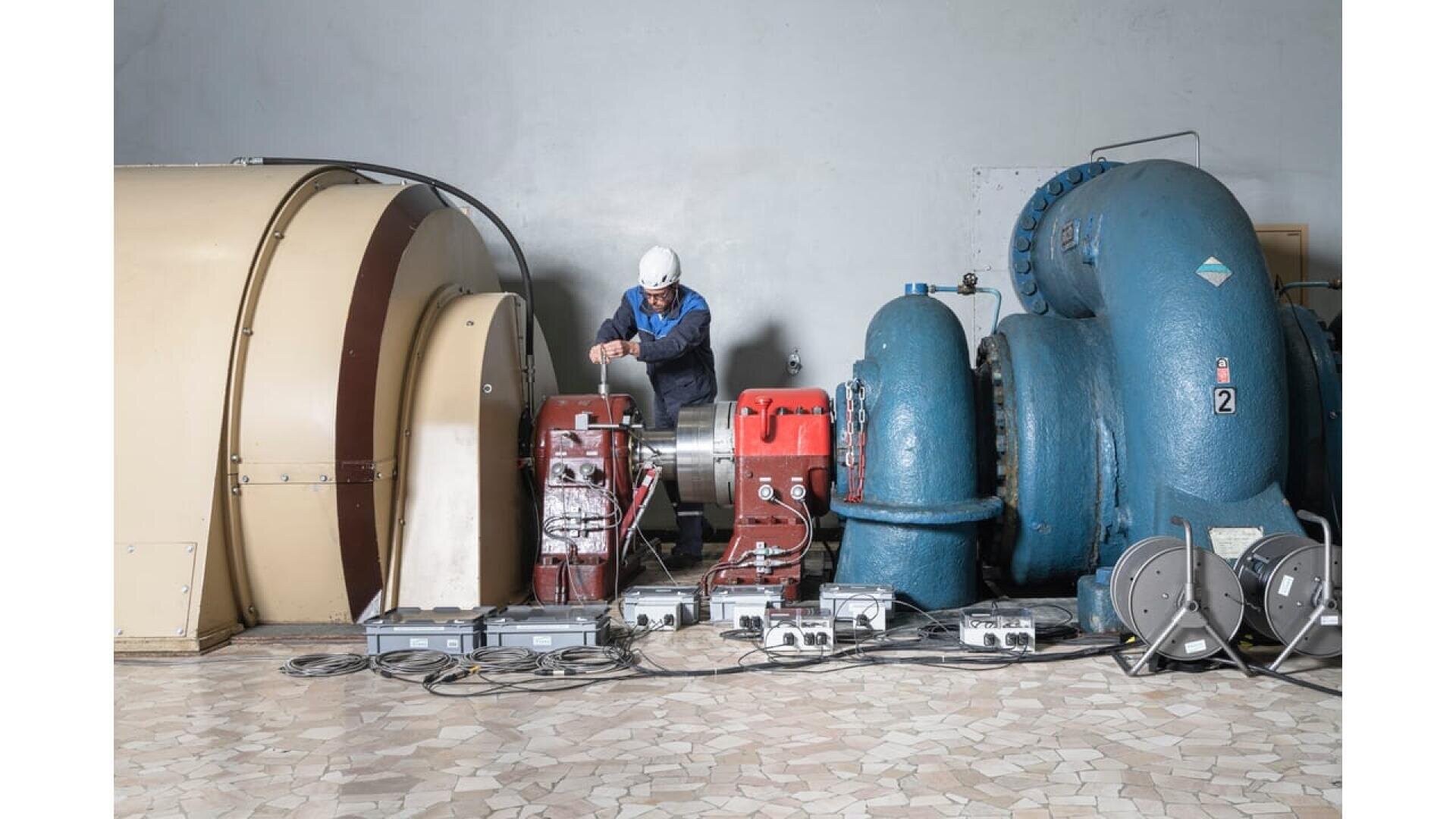 A worker in a white helmet operates machinery in an industrial room, surrounded by large blue and beige mechanical equipment and cables on a tiled floor.