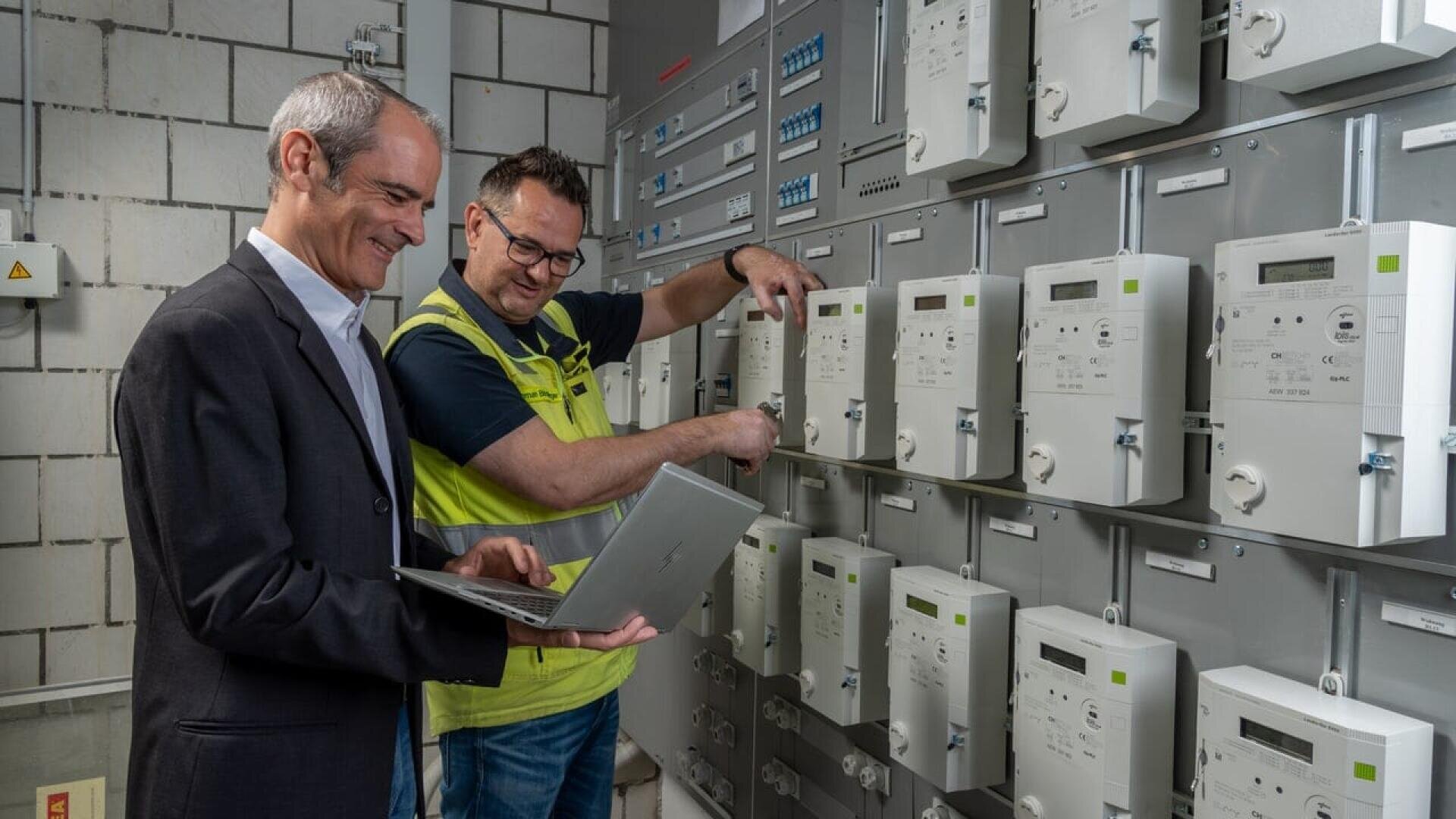 Two men work in a utility control room; one in a suit holds a laptop and smiles, while the other, wearing a vest, uses a screwdriver on a row of electrical meters mounted on the wall.