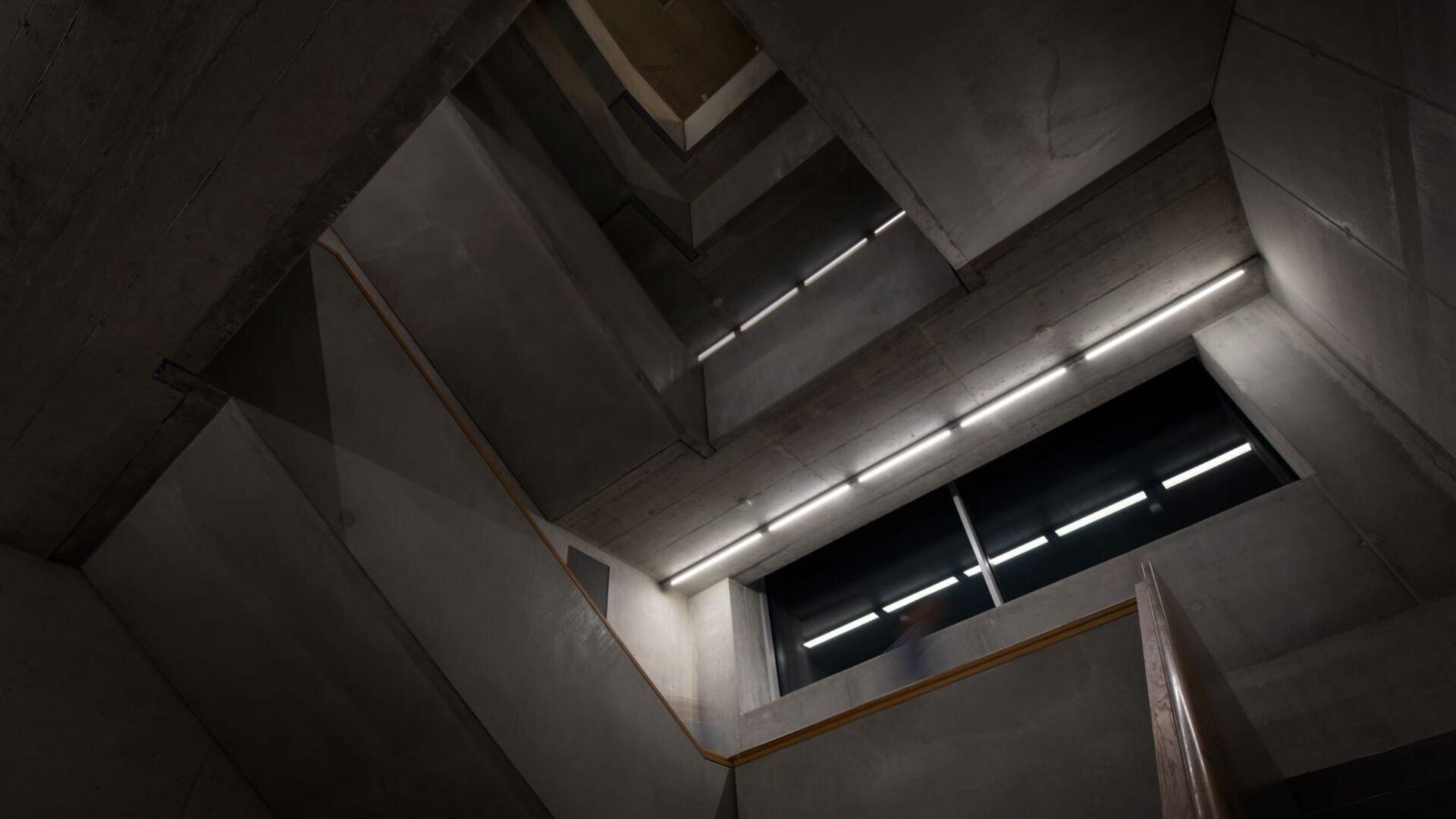Ein dramatischer, schräger Blick in ein modernes Betontreppenhaus mit geometrischen Linien, rechteckigen Fenstern, sichtbaren Balken und hellen Lichtbändern an der Decke und den Wänden.