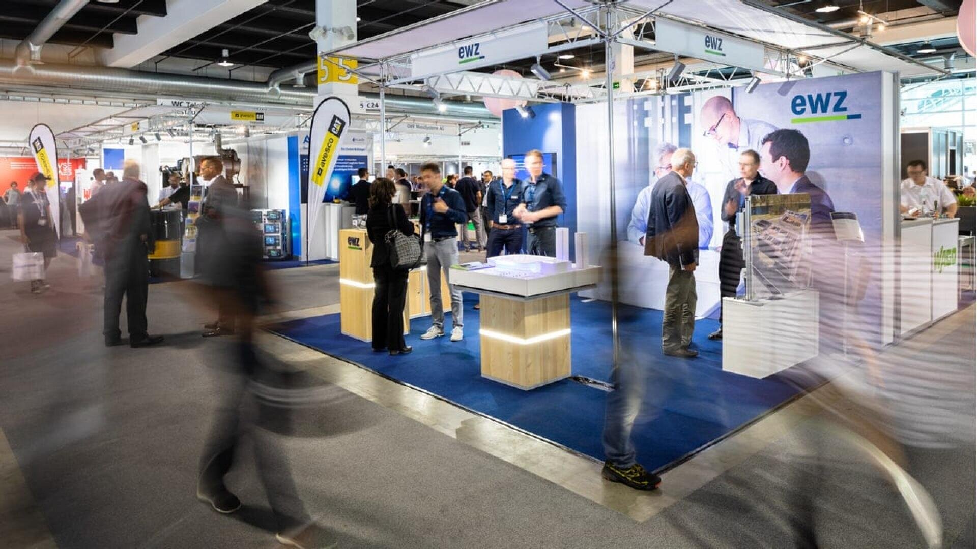 A busy trade show with people walking and chatting around an exhibition booth labeled ewz. The booth features displays, lighting, and promotional materials, with several attendees engaging in discussions.