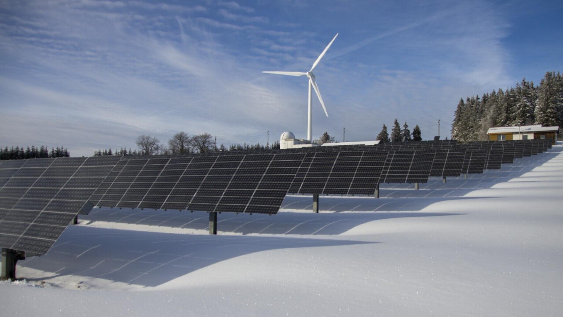 Reihen von Solarmodulen sind teilweise mit Schnee auf einem Feld bedeckt, mit einer großen Windturbine im Hintergrund. Schneebedeckter Boden und Bäume sind unter einem blauen Himmel mit Wolkenfetzen zu sehen.