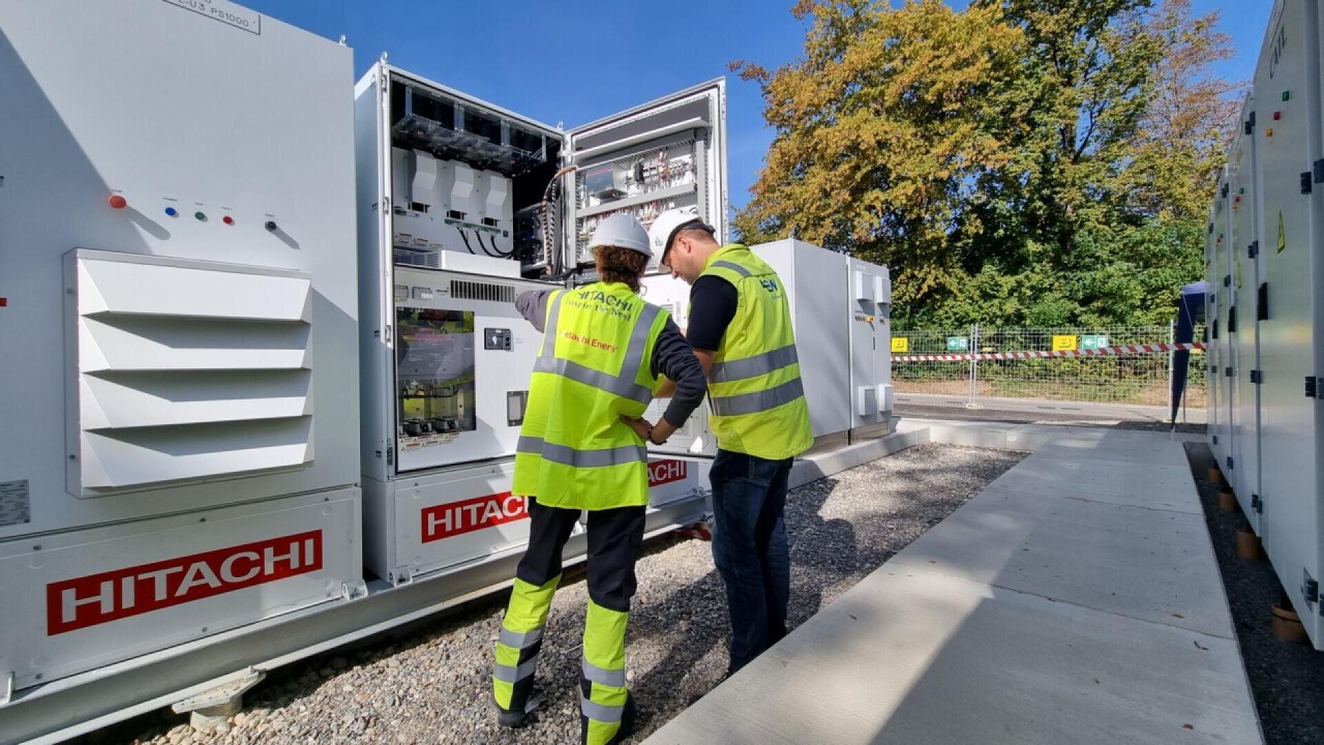 Two workers in safety vests and helmets inspect and discuss electrical equipment in an open Hitachi control cabinet outdoors, with trees and a clear sky in the background.