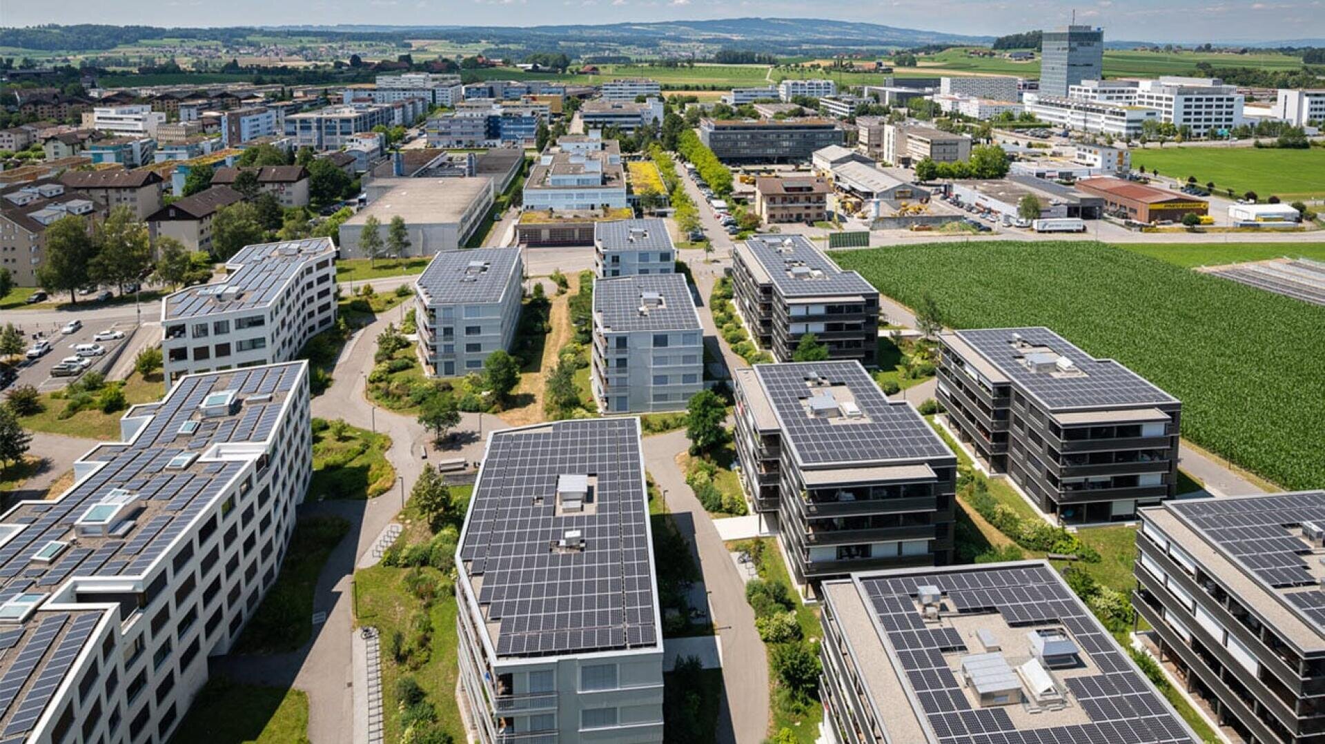 Luftaufnahme von modernen Wohn- und Geschäftsgebäuden mit Sonnenkollektoren auf den Dächern, umgeben von grünen Feldern und Straßen, unter einem klaren Himmel.