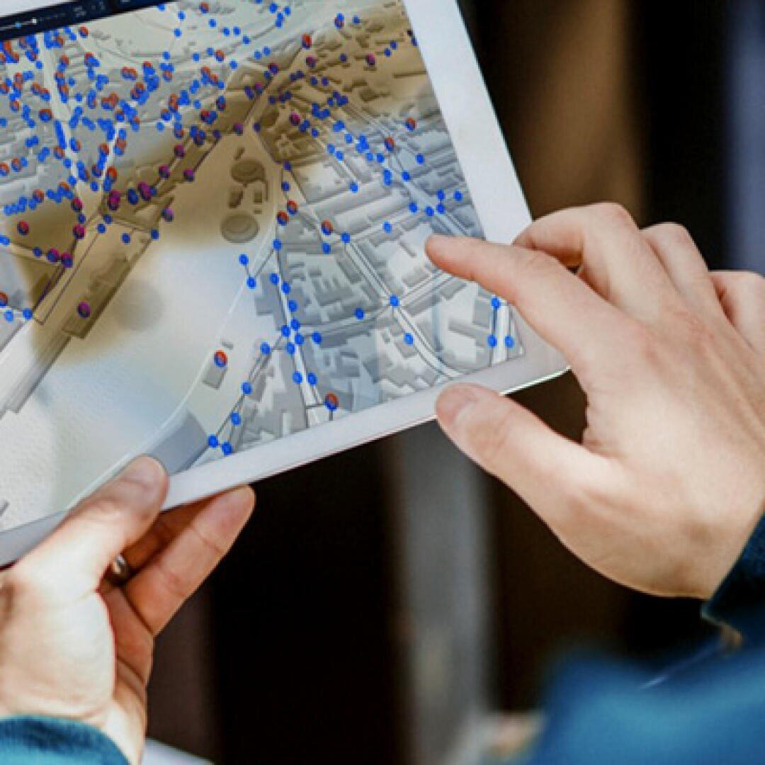 A person uses a tablet on which a digital map with numerous blue and red data points and a 3D layout of buildings is displayed.
