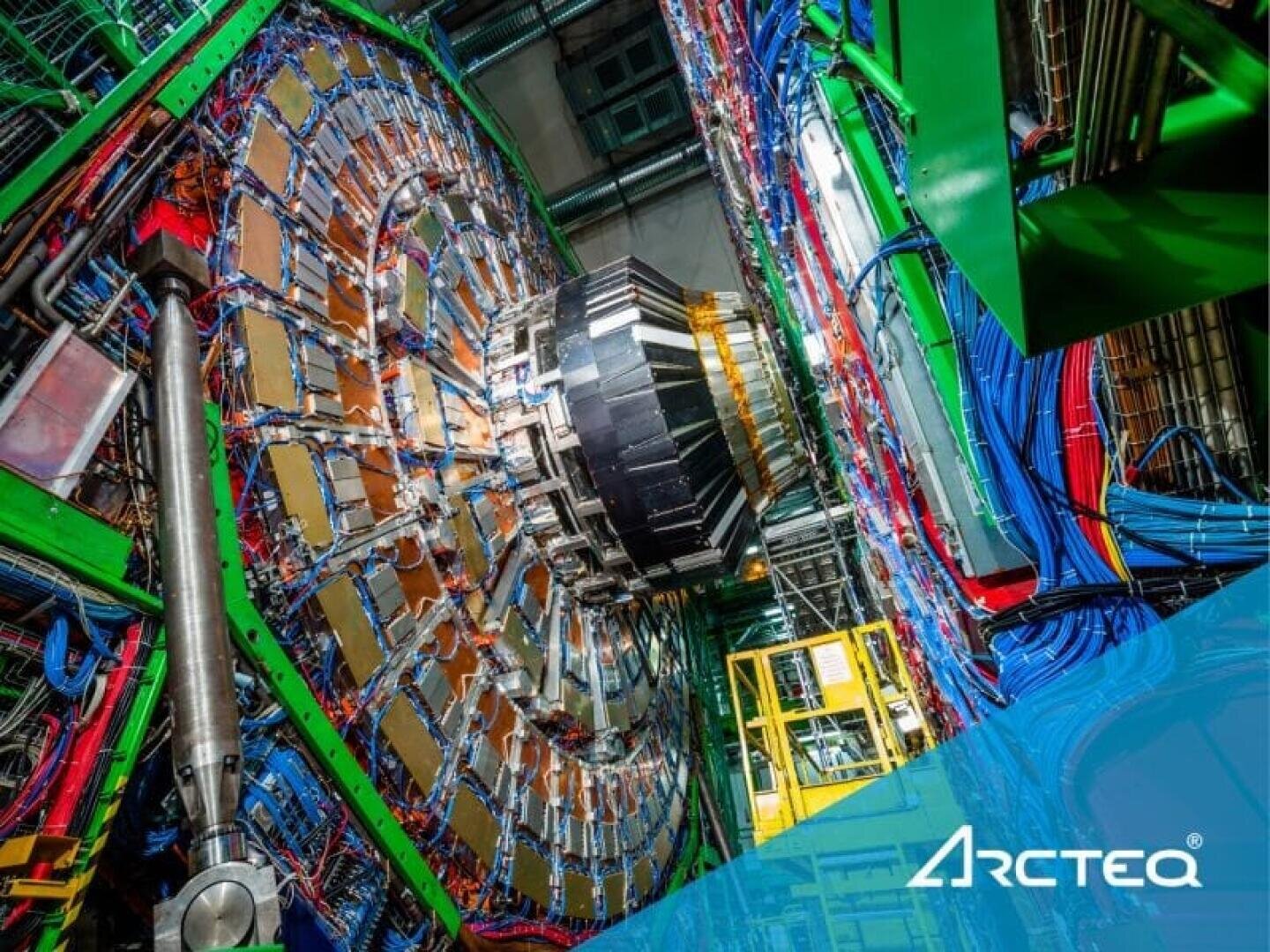 Ein großer, komplexer Teilchendetektor mit vielen bunten Kabeln und Metallkomponenten in einer Laborumgebung. Das Arcteq-Logo erscheint in der unteren rechten Ecke auf einem blauen Overlay.
