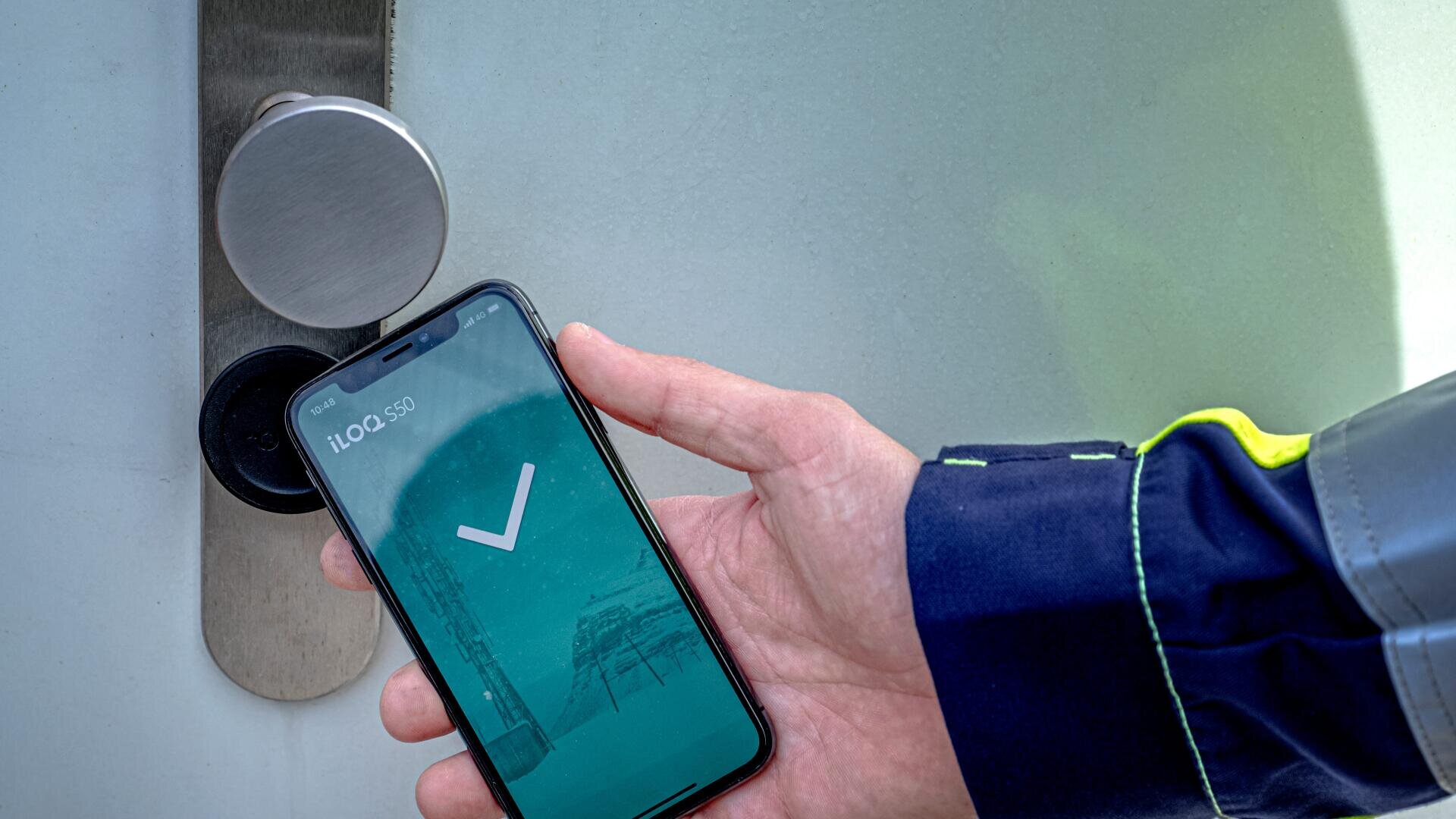 Eine Person hält ein Smartphone mit einem großen Häkchen auf dem Bildschirm in die Nähe eines digitalen Türschlosses und tut so, als würde sie die Tür mit dem Telefon entriegeln.