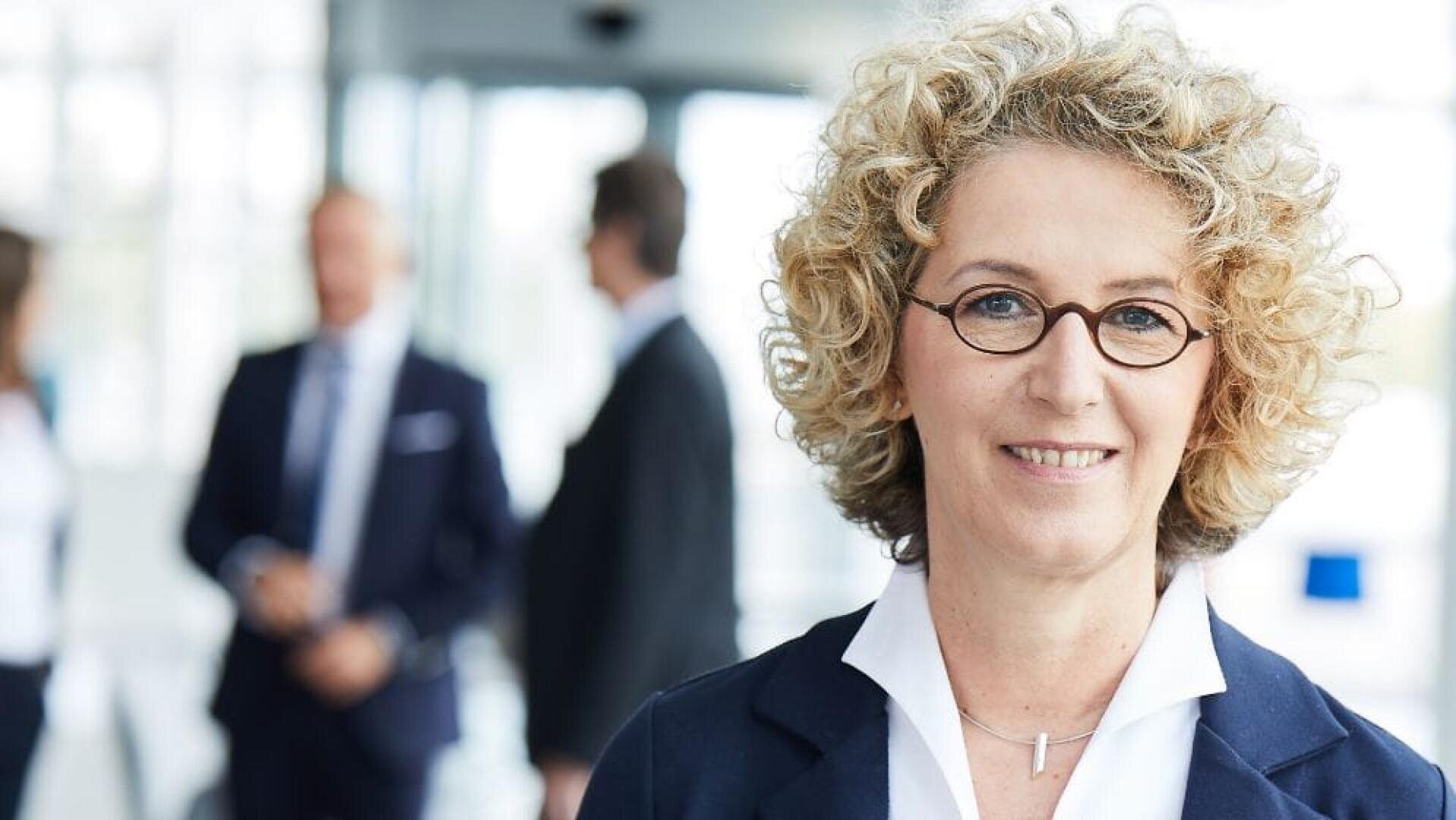 Eine lächelnde Frau mit kurzem, lockigem, blondem Haar und runder Brille steht im Mittelpunkt, sie trägt ein weißes Hemd und einen dunklen Blazer. Drei unscharfe Personen in Geschäftskleidung unterhalten sich im hellen Bürohintergrund.