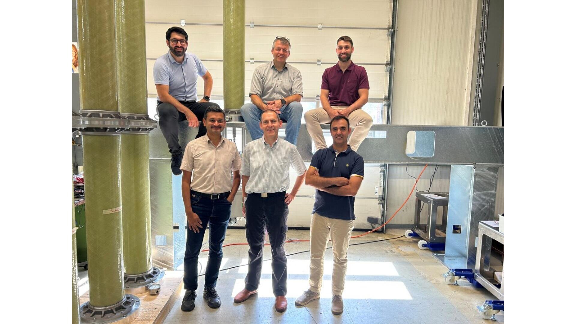 Six men pose and smile in an industrial-looking room with large green pillars and metal equipment. Three stand in front while three sit or perch on elevated surfaces behind them. Sunlight streams through a large window.