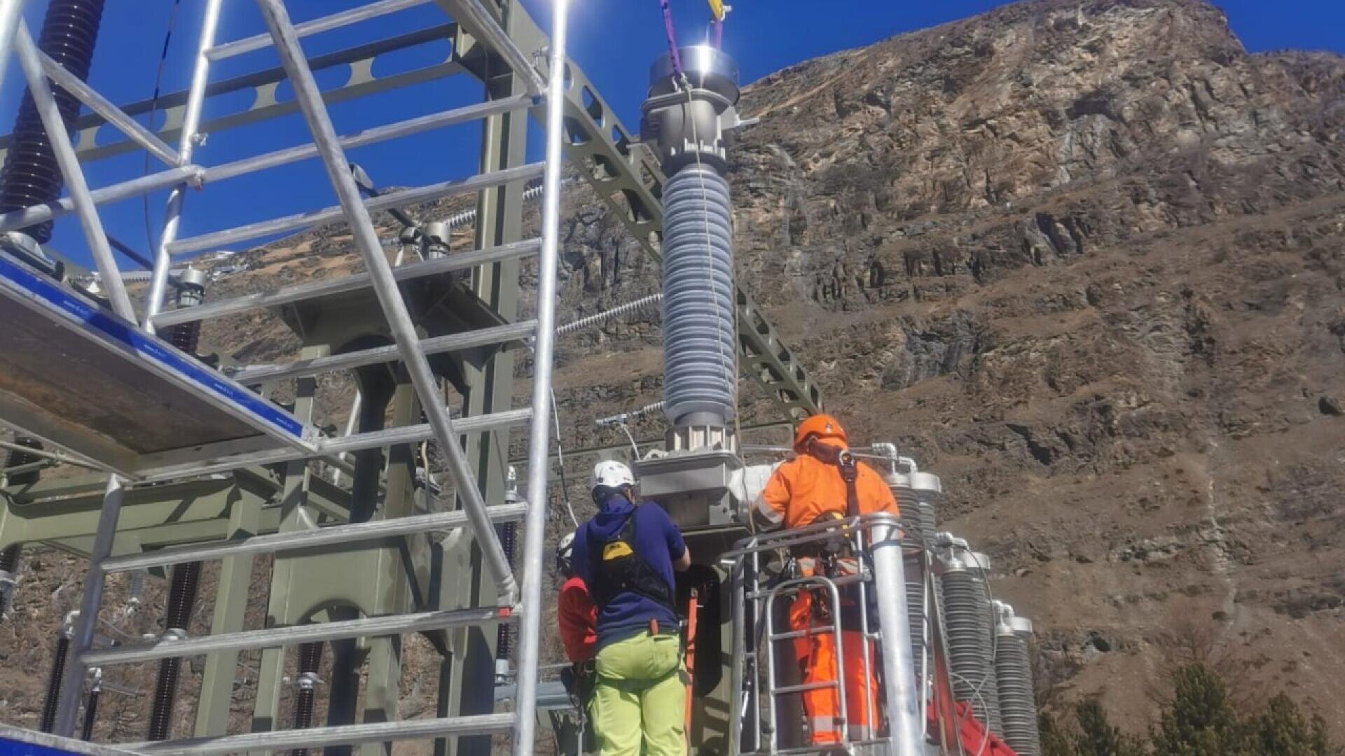 Two workers in safety gear inspect or repair electrical equipment on a metal structure outdoors, with rocky mountains visible in the background under a clear blue sky.