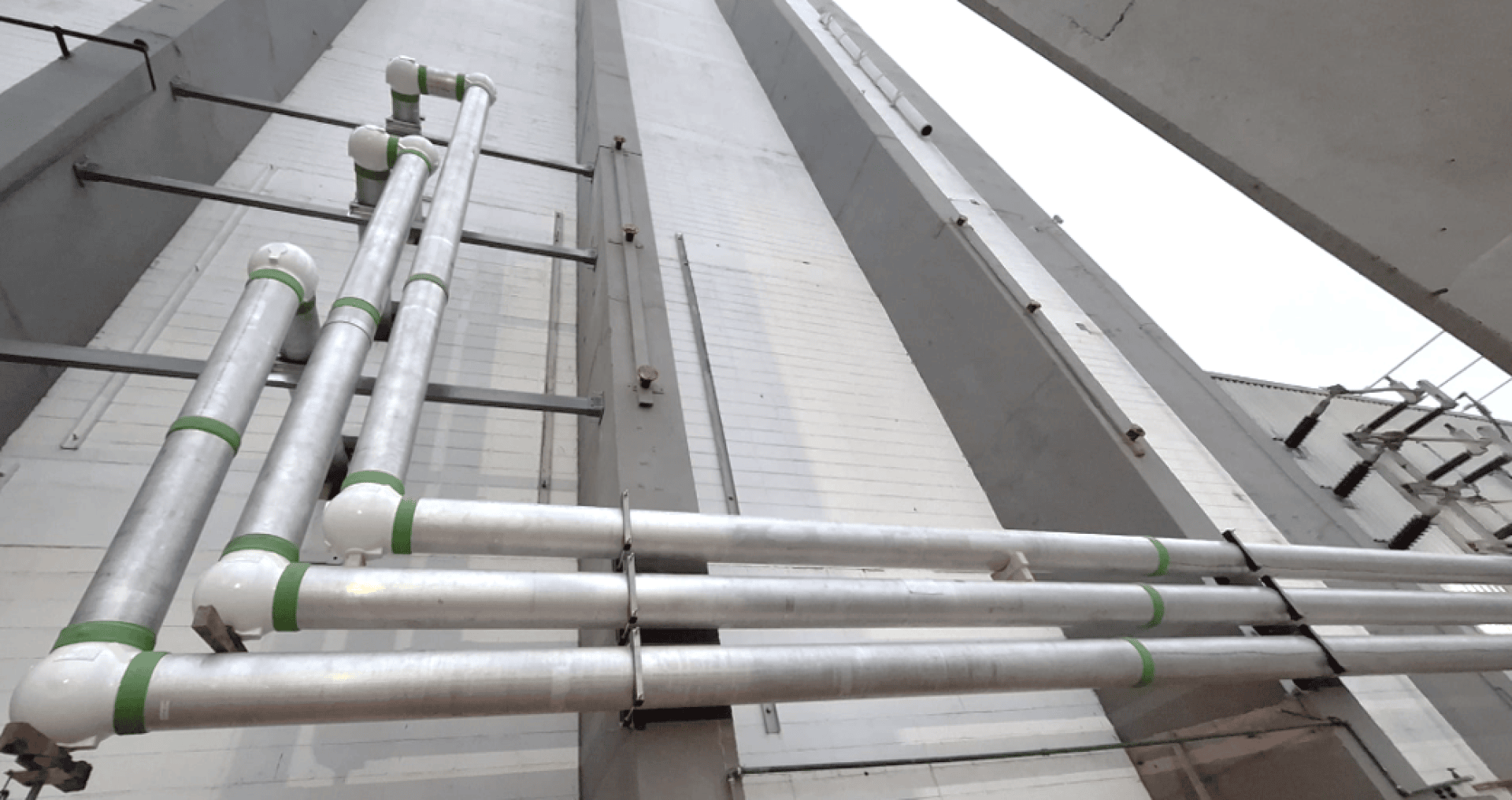 Three parallel silver metal tubes with green markings run vertically and horizontally along the outer wall of a tall building and are held in place by metal brackets. The photo is taken from a low angle, looking upwards.