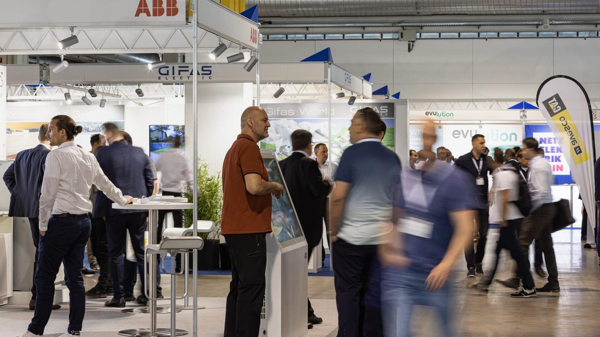 A busy indoor trade show with several people standing, talking, and walking past exhibition booths displaying technology and business banners. Some attendees are blurred, indicating movement.