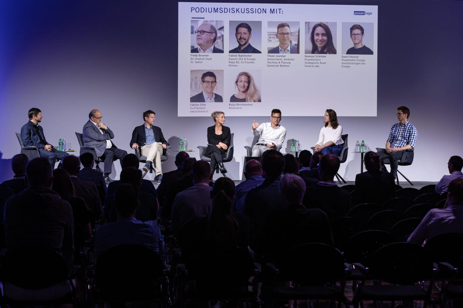 Sieben Personen sitzen bei einer Podiumsdiskussion auf der Bühne, ein Publikum schaut zu. Hinter ihnen wird eine Leinwand projiziert, auf der unter der Überschrift PODIUMSDISKUSSION MIT die Namen der Diskussionsteilnehmer zu sehen sind.