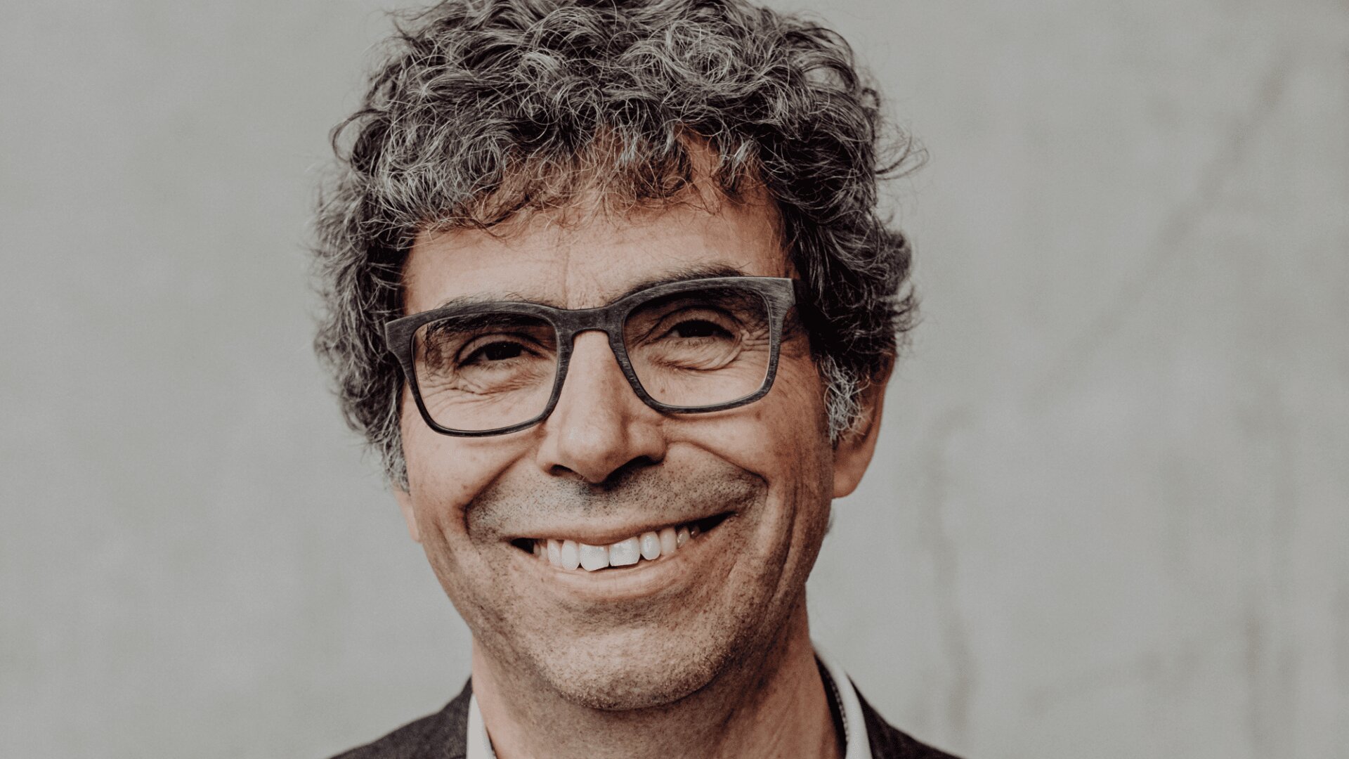 Ein Mann mittleren Alters mit lockigem grauem Haar, einer dunklen rechteckigen Brille und einem leichten Lächeln steht vor einem schlichten, hellen Hintergrund.