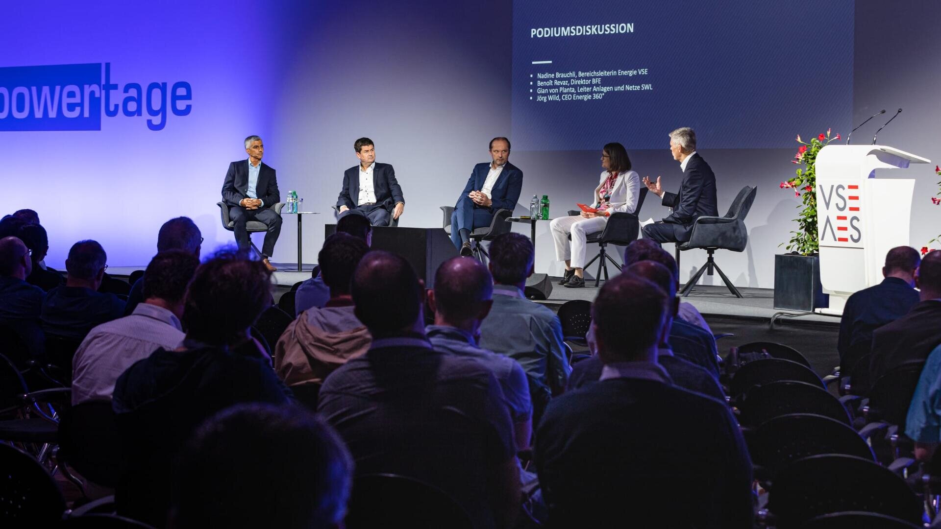 A panel of five people in business attire sit on stage discussing in front of an audience at a conference. A presentation slide behind them reads PODIUMSDISKUSSION. The room is dimly lit except for the stage.