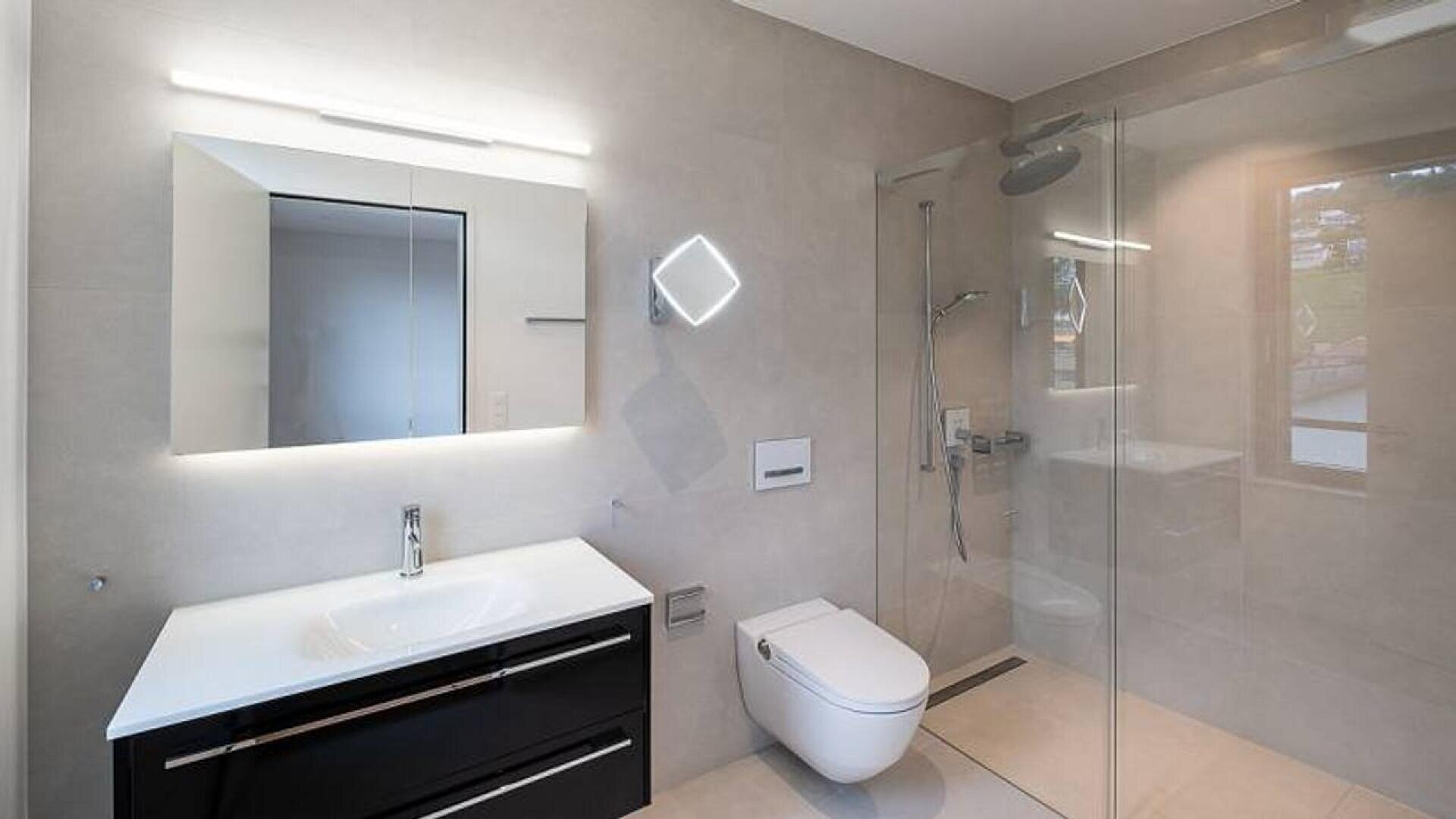 Modern bathroom with a wall-mounted sink and cabinet, illuminated mirror, floating toilet, and a glass-enclosed shower with a rain showerhead; neutral light gray tiles cover the walls and floor.
