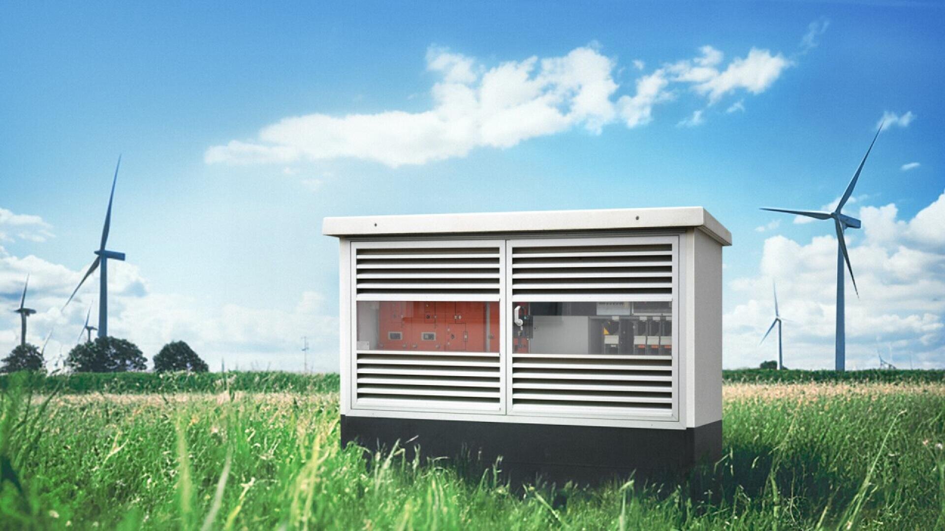 A white electrical utility box stands in a grassy field, with wind turbines and trees in the background under a bright blue sky with scattered clouds.