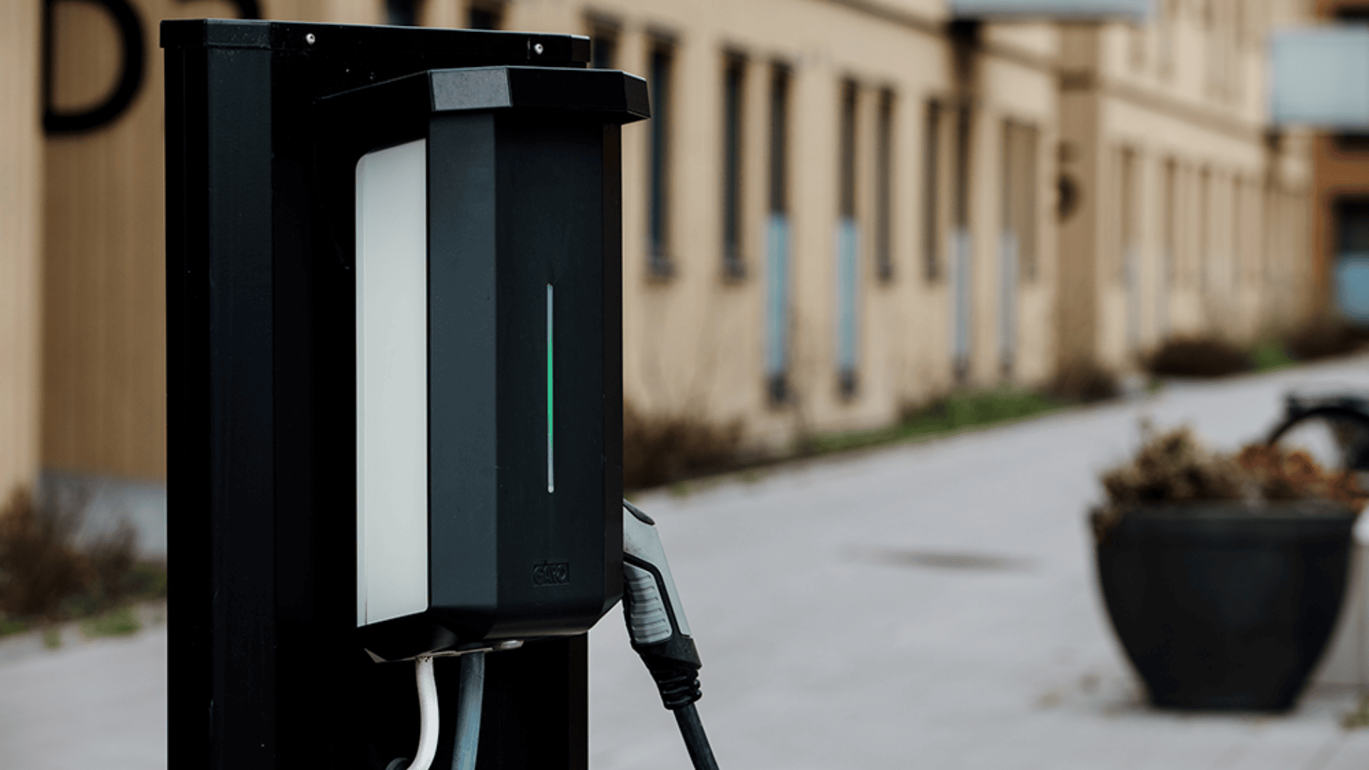 Eine Nahaufnahme einer Ladestation für Elektrofahrzeuge mit eingestecktem Kabel, die sich im Freien in der Nähe eines Gebäudes und eines großen Pflanzkübels auf einem gepflasterten Weg befindet.