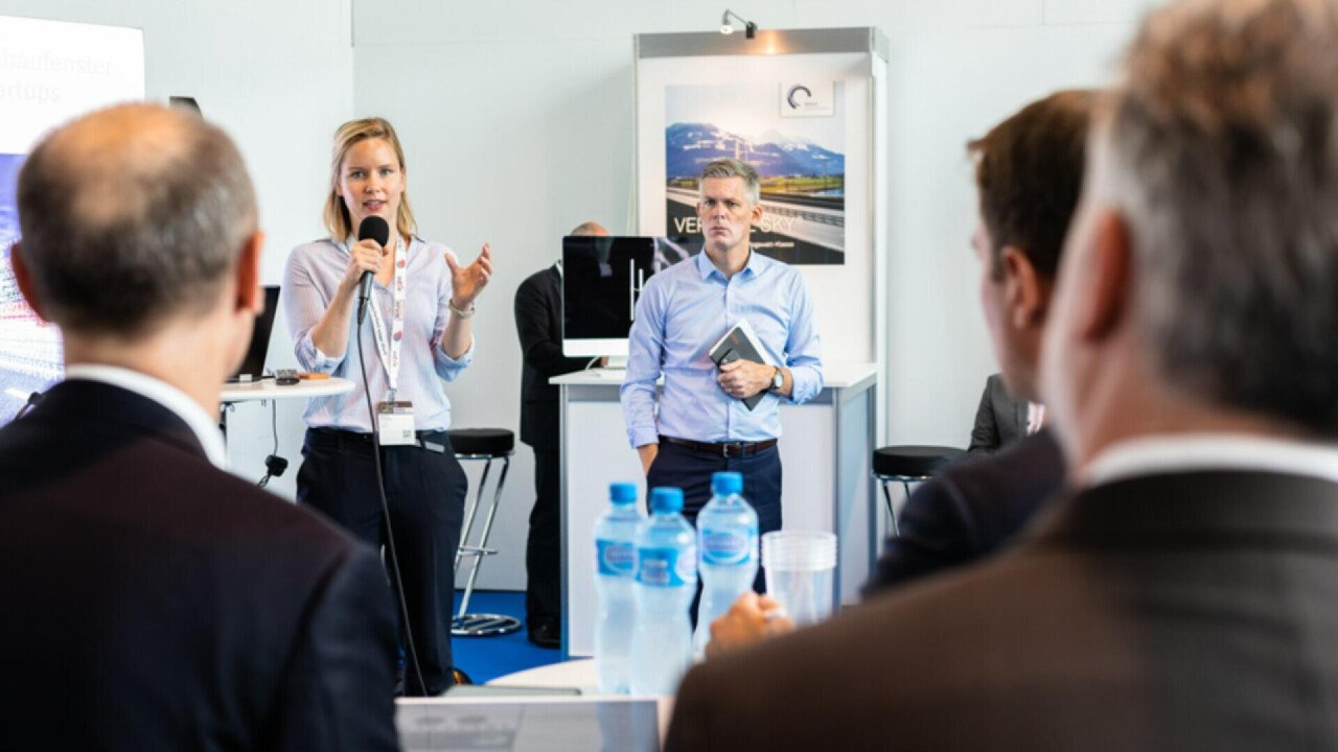 A woman is speaking into a microphone in front of an audience at a business event. A man stands nearby holding a notebook, while several people listen attentively. Bottles of water are visible on a table in the foreground.