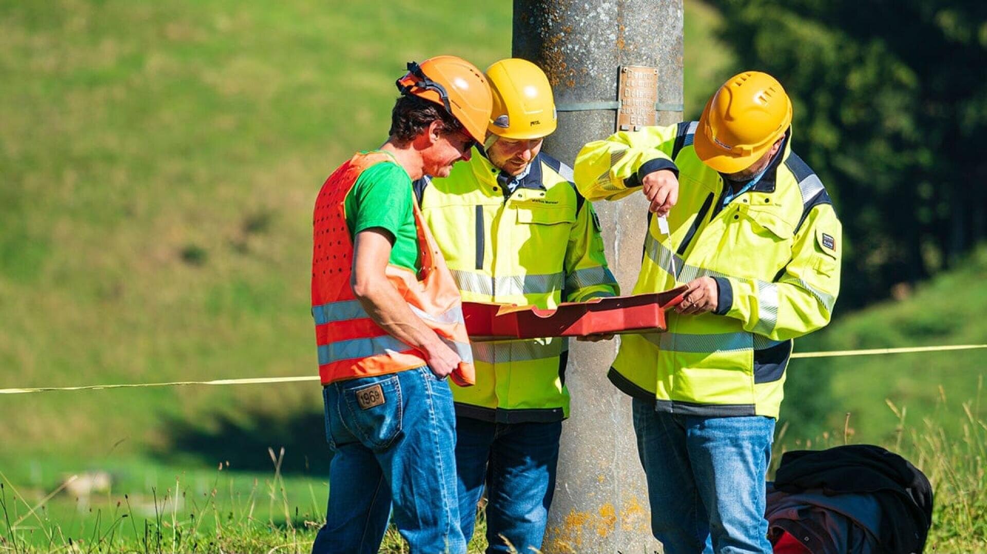 Drei Arbeiter in Sicherheitsausrüstung stehen in der Nähe eines Mastes im Freien. Zwei, die gelbe Jacken und Helme tragen, prüfen Papiere auf einem Klemmbrett, während der dritte, der ein grünes Hemd und eine orangefarbene Weste trägt, zuschaut. Im Hintergrund sind Gras und Hügel zu sehen.