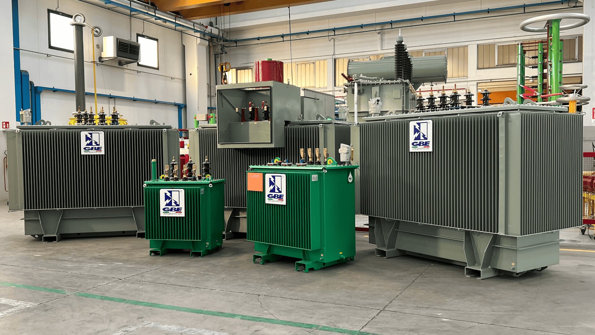 Several industrial transformers of varying sizes, mostly gray and one green, are displayed inside a warehouse with concrete floors and high ceilings. Logos with CBE are visible on the equipment.