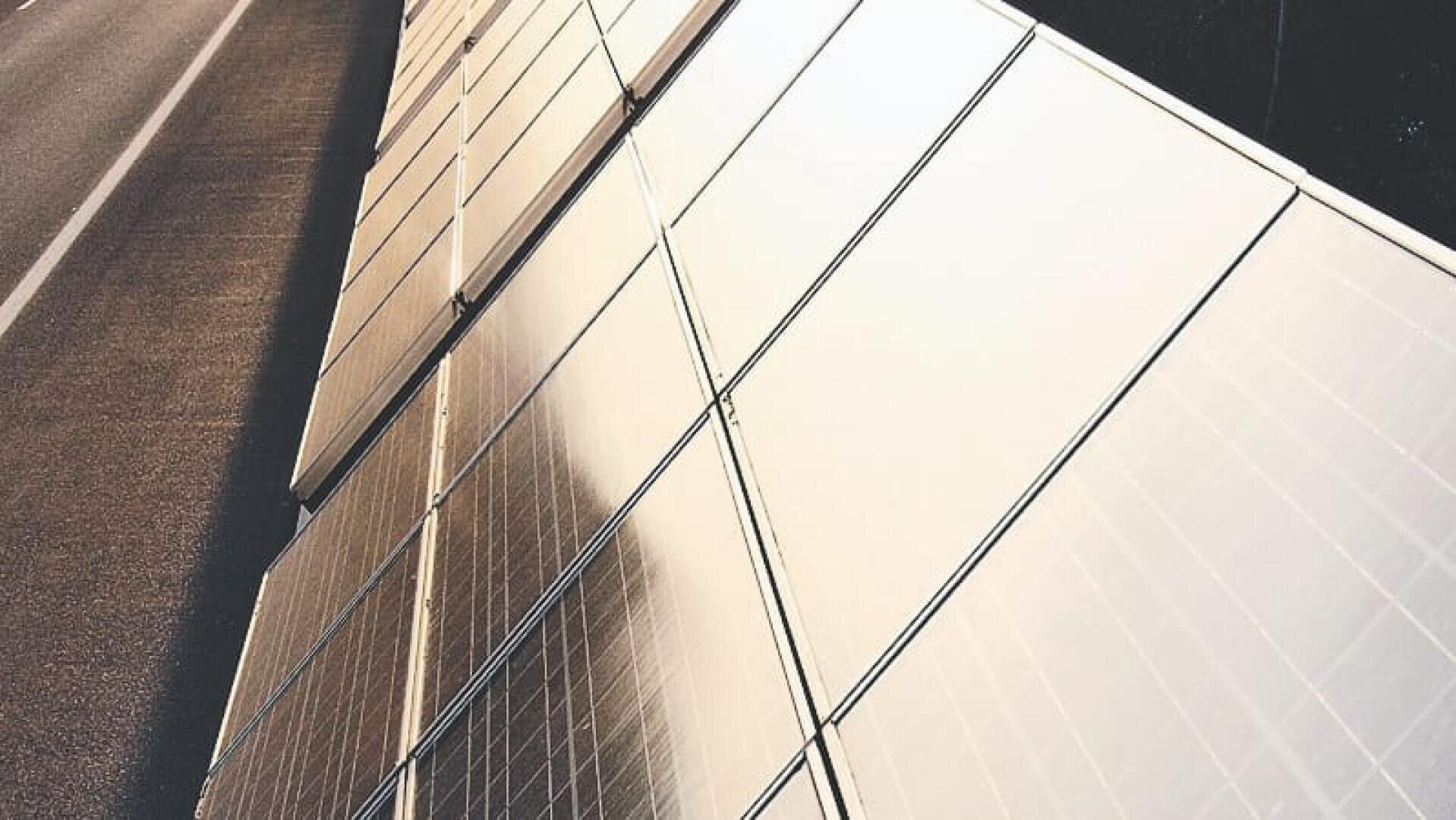 Reihen von Solarmodulen sind schräg neben einer Straße angebracht, reflektieren das Sonnenlicht und erzeugen erneuerbare Energie. Die Paneele erzeugen bei hellem Licht ein geometrisches Muster.