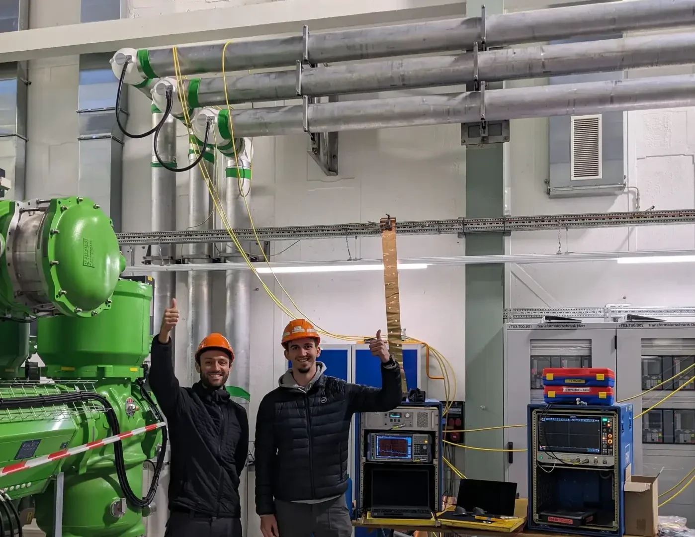 Two people wearing orange hard hats and black jackets stand in an industrial laboratory, smiling and giving a thumbs-up. One of them is holding a long copper strip; equipment, cables and machines can be seen in the background.