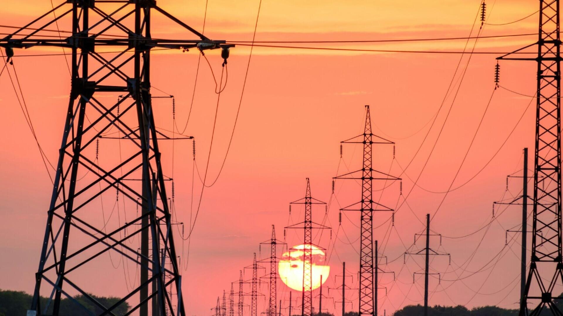 High-voltage power lines and transmission towers stretch across the landscape at sunset, silhouetted against a vibrant orange and pink sky with the sun low on the horizon.