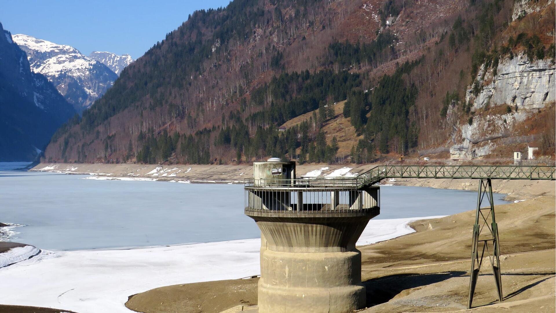 Ein Wasserentnahmeturm aus Beton steht auf trockenem Boden neben einem teilweise zugefrorenen Stausee, umgeben von steilen, bewaldeten Bergen mit Schneeflecken unter einem klaren blauen Himmel.
