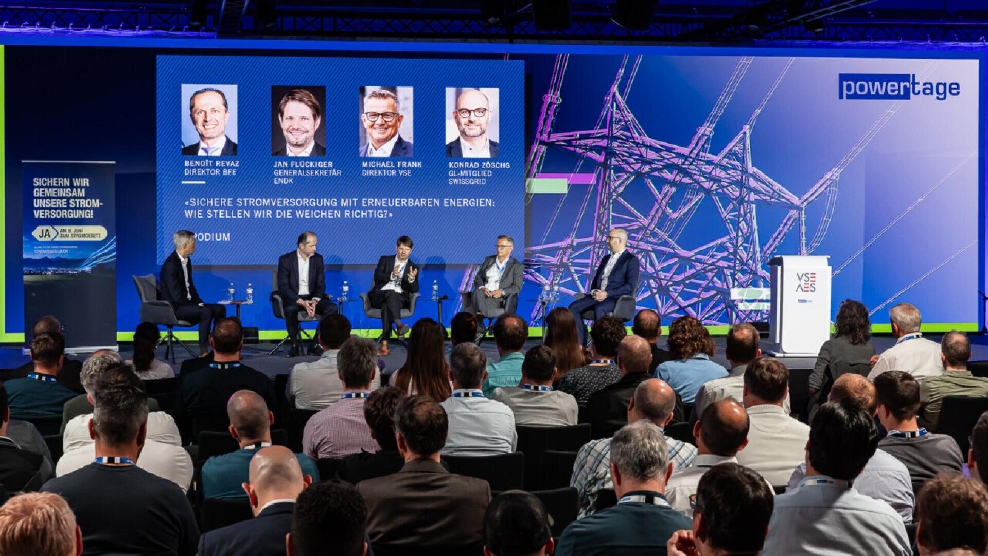 A panel of five men in business attire sit on stage discussing energy at a conference, with an audience seated in front. A large screen behind them displays their names, photos, and the conference topic on renewable energy.