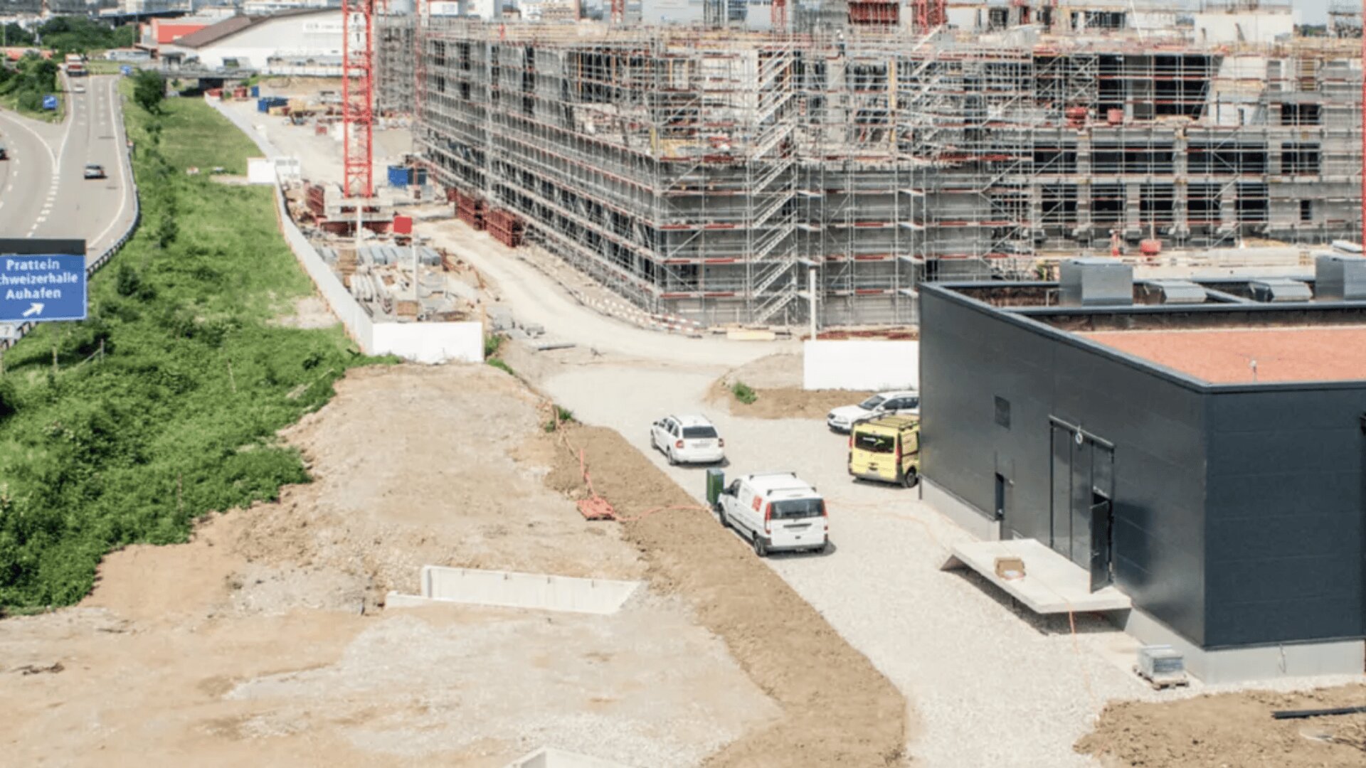 Luftaufnahme einer Baustelle mit Gerüsten an einem großen Gebäude, auf einer Schotterstraße geparkten Fahrzeugen und einem schwarzen Gebäude im Vordergrund neben einer teilweise gepflasterten Fläche. Links sind eine Straße und Grünflächen zu sehen.