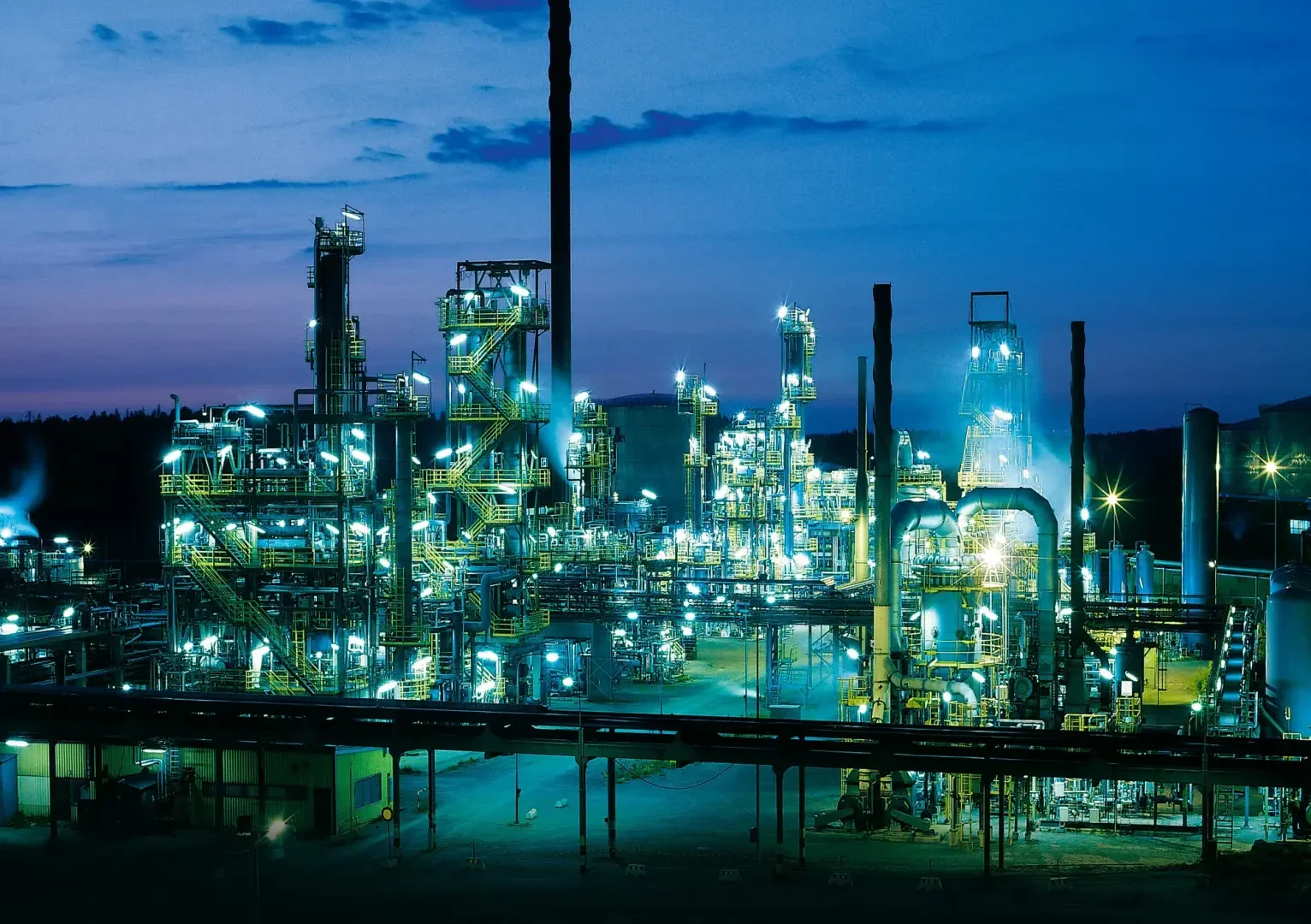 Ein beleuchteter industrieller Raffineriekomplex in der Abenddämmerung, mit hohen Metallstrukturen, Rohren und Lichtern, die vor dem blauen Himmel leuchten.