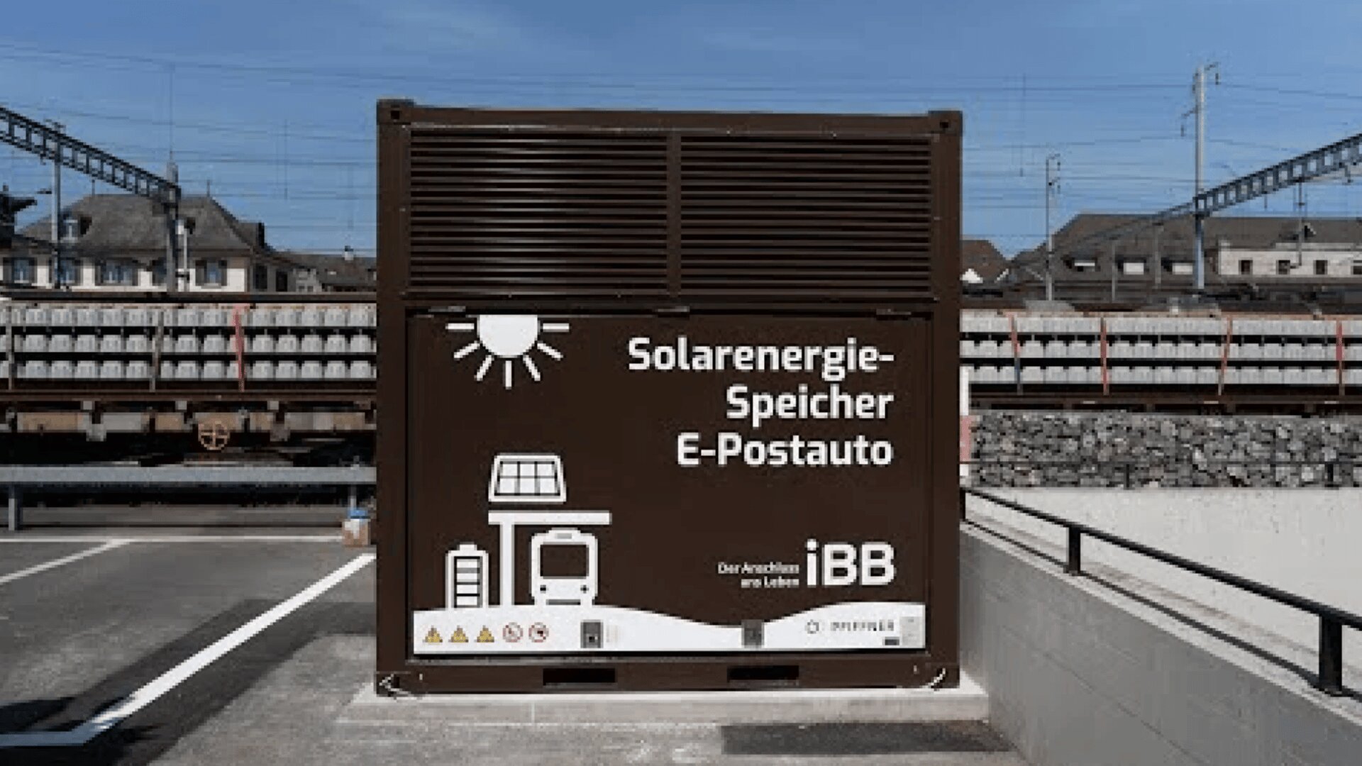 Großer brauner Container mit der Aufschrift "Solarenergie-Speicher E-Postauto" und weißen Symbolen, die Solarzellen, einen Bus und das Aufladen darstellen. Er steht im Freien in der Nähe von Bahngleisen und Gebäuden unter freiem Himmel.
