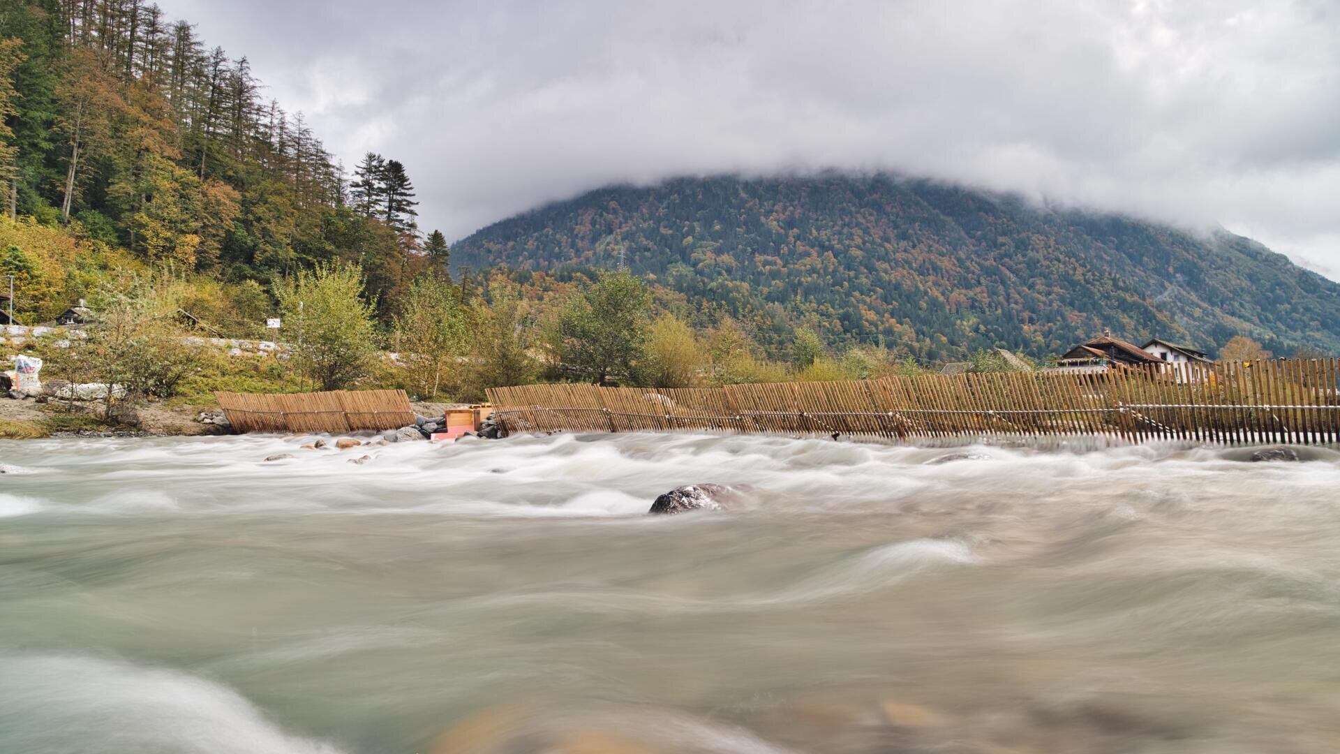 Ein schnell fließender Fluss mit glattem, verschwommenem Wasser fließt an Holzzäunen vorbei, umgeben von grünen Bäumen und Bergen, die an einem bewölkten Tag mit Nebel und niedrigen Wolken bedeckt sind.
