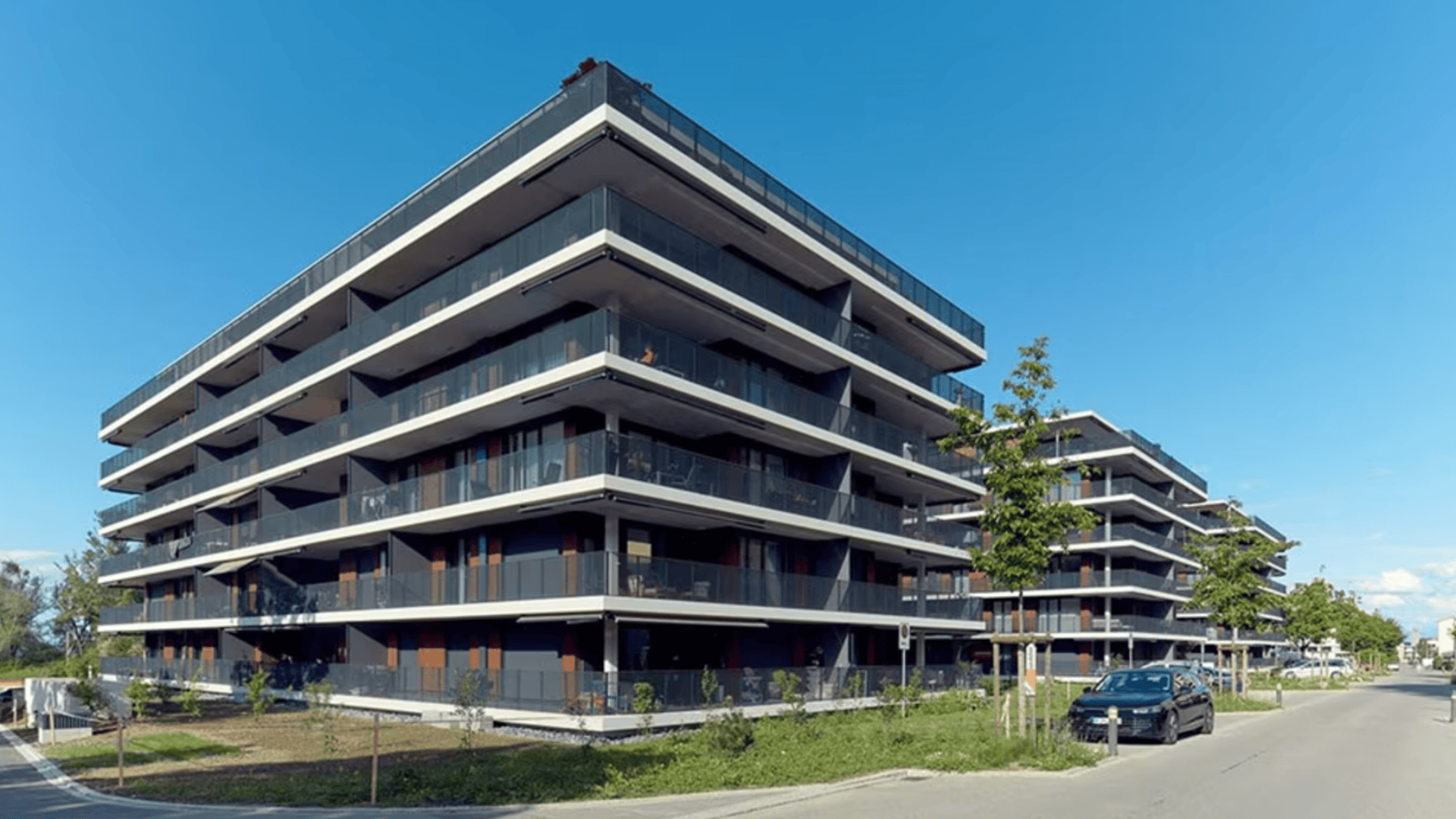 Moderne Mehrfamilienhäuser mit dunklen Glasbalkonen und Flachdächern, entlang einer ruhigen Straße mit parkenden Autos und jungen Bäumen unter einem klaren blauen Himmel.