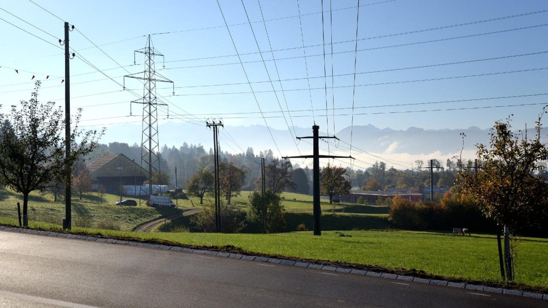 Eine ländliche Landschaft mit grünen Feldern, vereinzelten Bäumen und Stromleitungen, die sich über dem Boden erstrecken. In der Mitte des Bildes befinden sich eine Scheune und ein Bauernhaus. Im Hintergrund sind die Berge und der Morgennebel unter einem klaren blauen Himmel zu sehen.