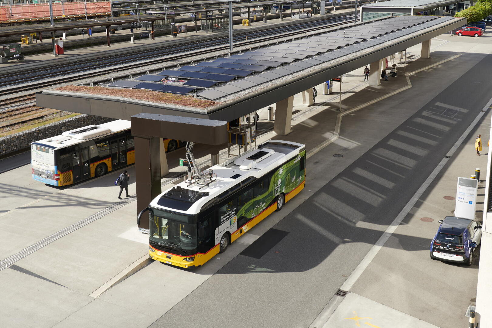 Ein gelber und grüner Elektrobus lädt an einer mit Solarzellen bedeckten Station neben den Bahngleisen auf; ein weiterer Bus wartet in der Nähe. Menschen gehen auf dem Bahnsteig und einige Autos sind neben dem Bahnhof geparkt.