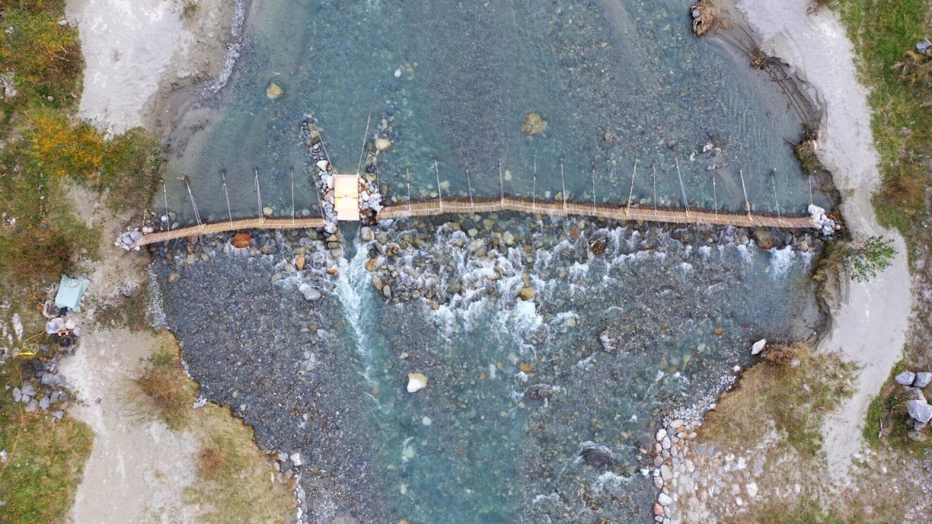 Aerial view of a narrow wooden footbridge crossing a shallow, rocky river with clear blue water, surrounded by patches of grass and sand on both banks.
