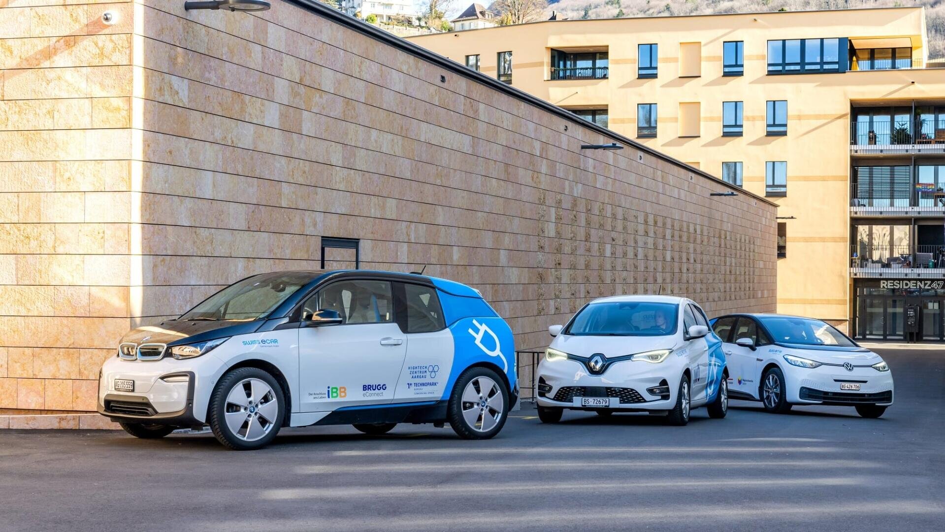 Drei Elektroautos sind in einer Reihe entlang eines modernen Gebäudes geparkt. Die Autos sind mit blauen und weißen Grafiken versehen, und die Umgebung scheint ein städtisches Gebiet mit Wohnungen im Hintergrund zu sein.