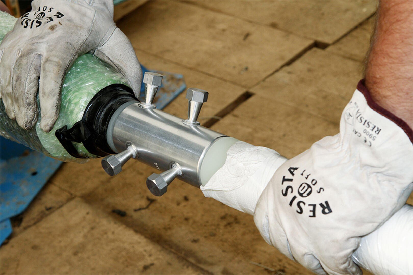 Two gloved hands are connecting two pipes using a metal coupling with bolts on a wooden surface. The gloves are labeled Resista Soft.