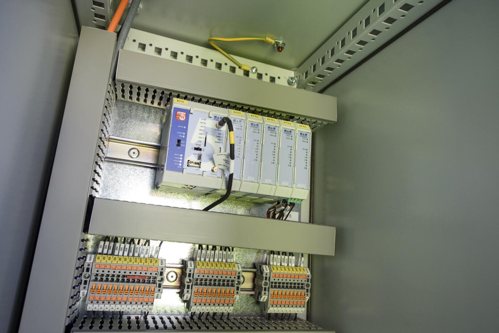 Eine elektrische Schalttafel mit Schaltmodulen, Kabeln und Klemmenblöcken, die übersichtlich in einem Metallgehäuse angeordnet sind. Ein gelbes Kabel ist mit einem Erdungspunkt oben rechts verbunden.