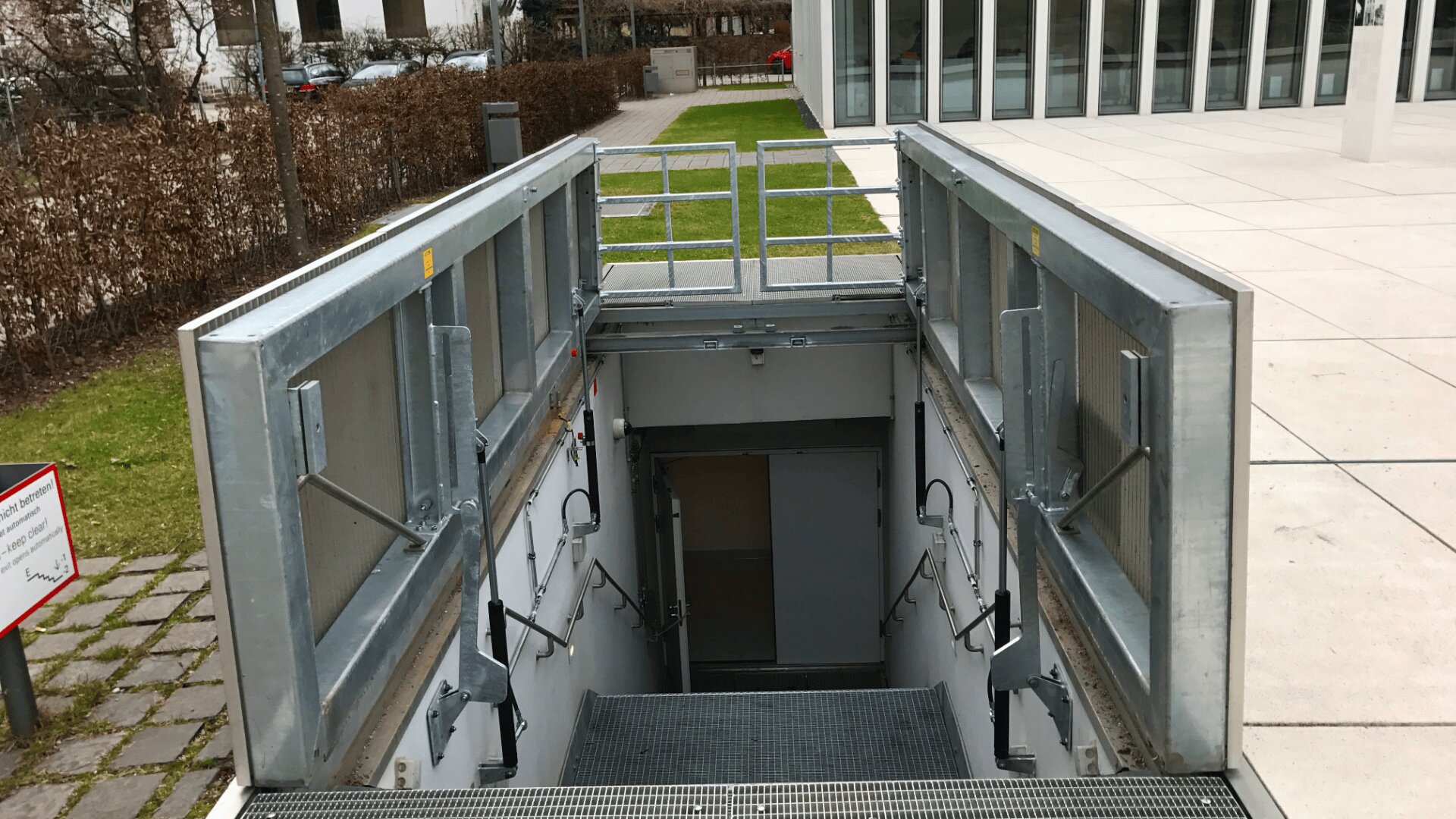 Eine moderne Treppe mit Metallrahmen führt hinunter zu einem unterirdischen Eingang außerhalb eines Gebäudes mit großen Glasfenstern und einem Betonweg. Gras und Hecken begrenzen das Gelände.